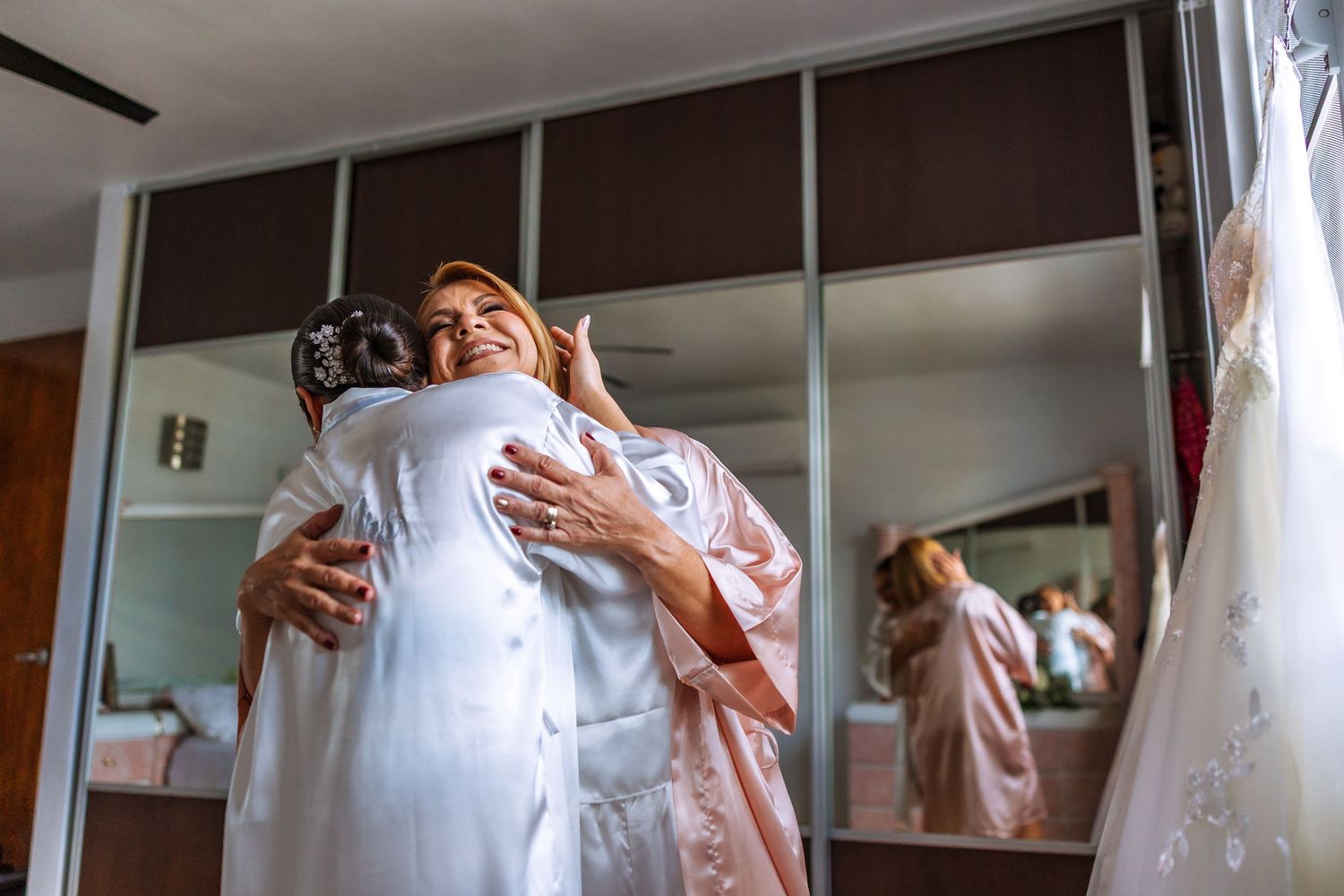 Two women in robes share a warm hug in a bridal suite with a wedding dress hanging nearby.