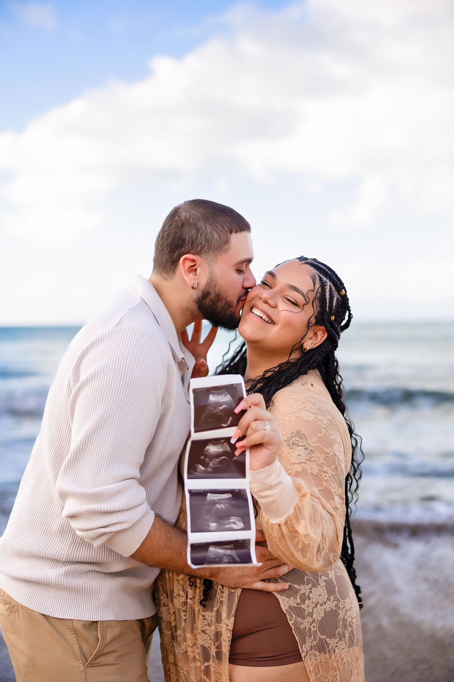 Couple holding ultrasound photos on beach, man kissing woman's cheek as she smiles, pregnancy announcement photo.