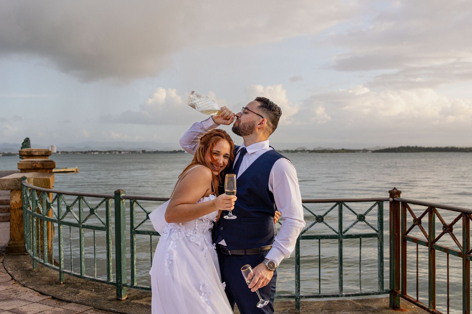 Bride and groom celebrating with champagne on a waterfront terrace at sunset on their wedding day.