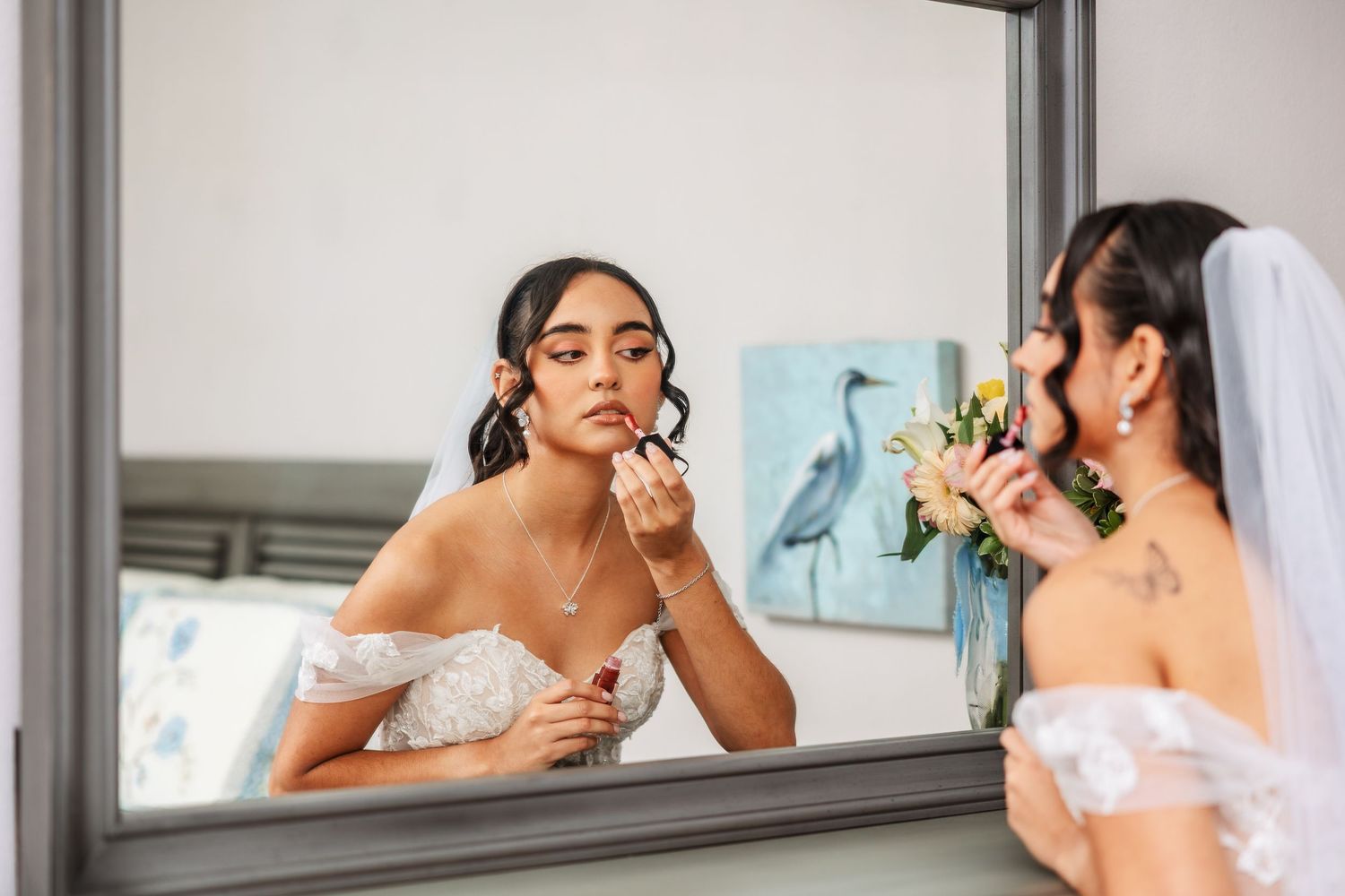 Bride in white off-shoulder gown applies makeup in mirror, holding floral bouquet, with veil and tattoos visible.