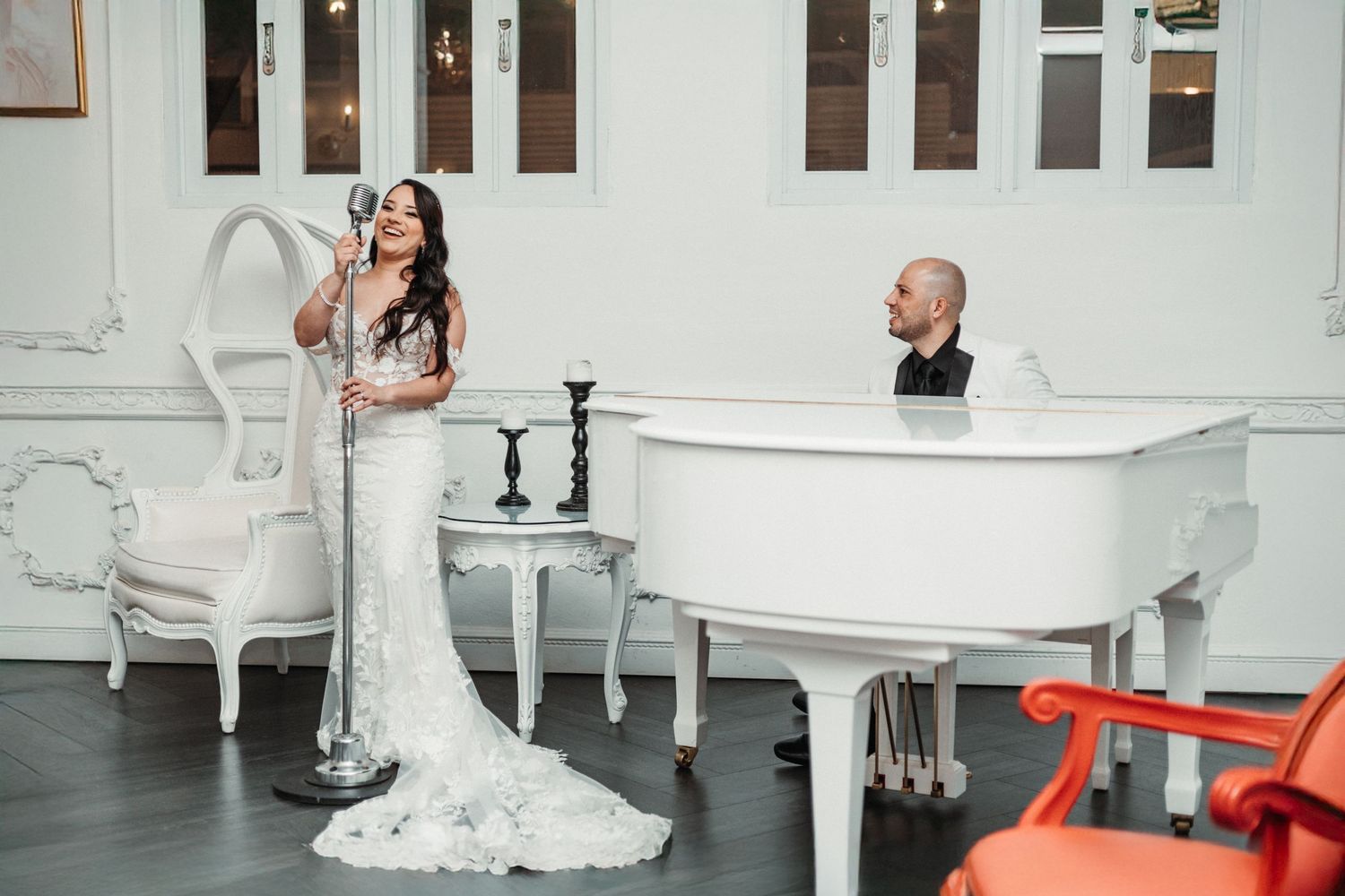 Bride in white gown singing at microphone while groom plays white grand piano indoors.