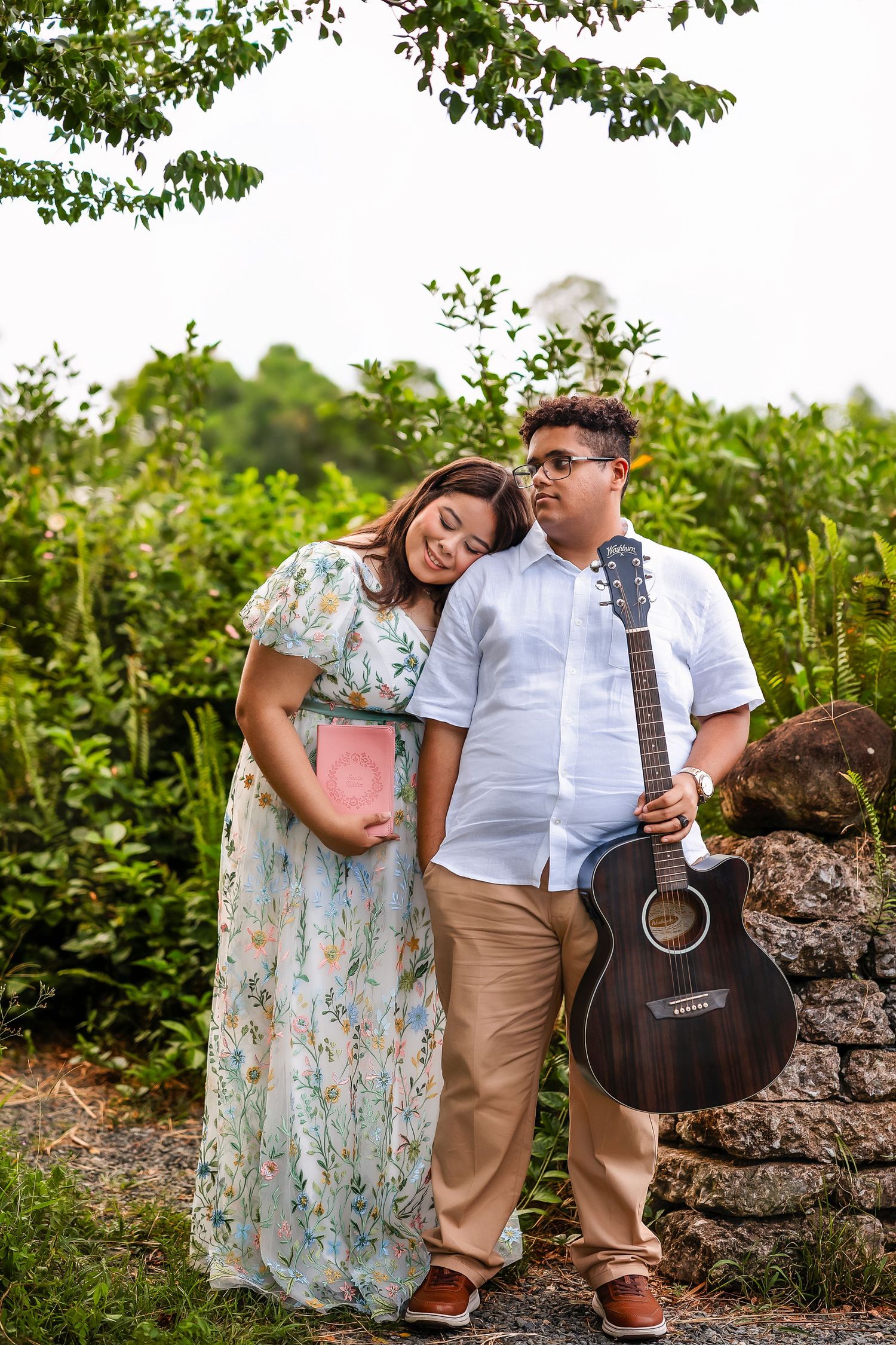 Pregnant woman in floral dress leans on man holding acoustic guitar outdoors near stone wall and lush greenery.