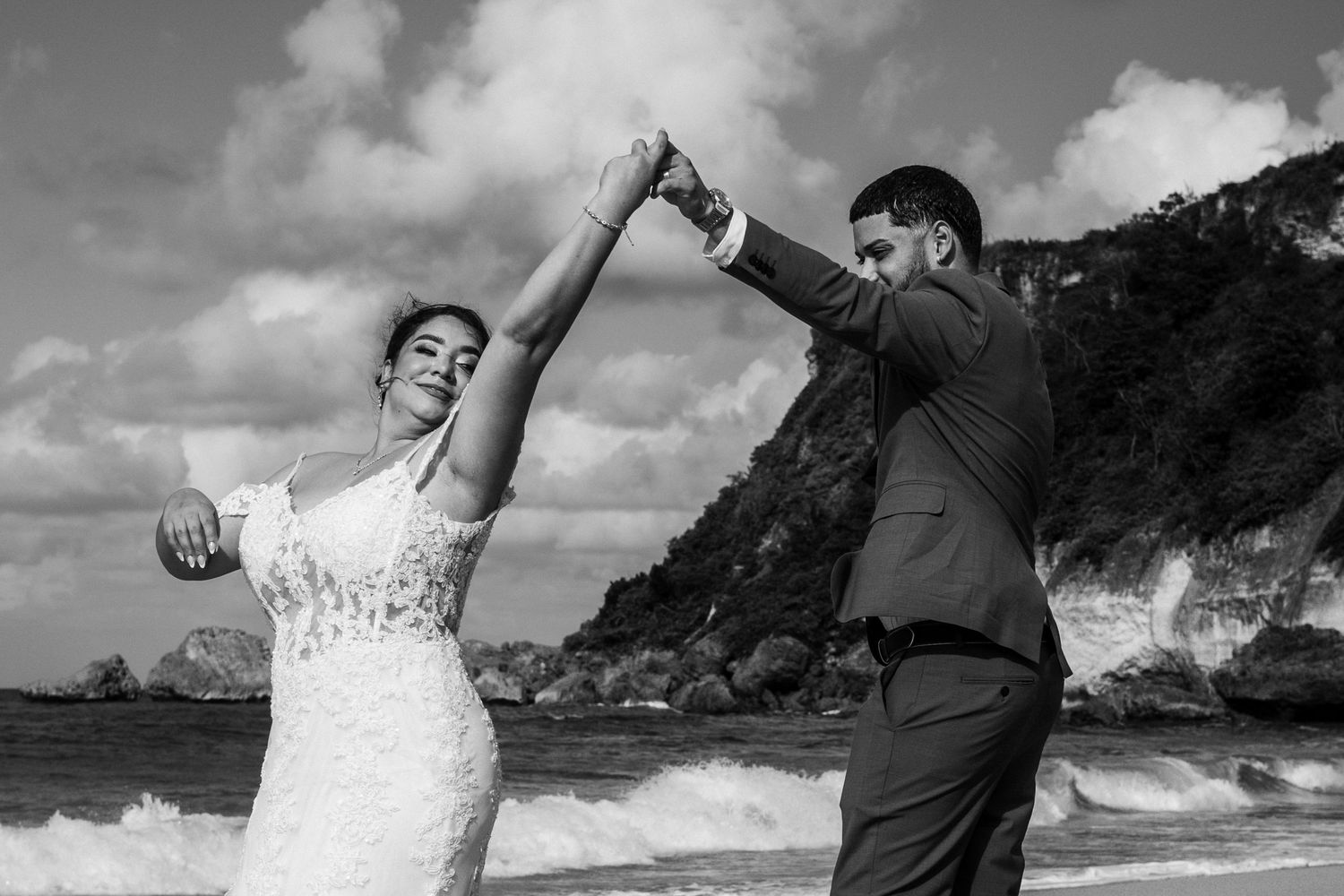 Dancing couple in wedding attire twirling on dramatic coastal clifftop in black and white.