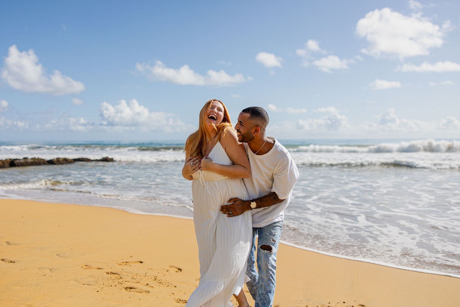 Happy couple laughing and embracing on a sunny tropical beach with blue ocean waves in the background.
