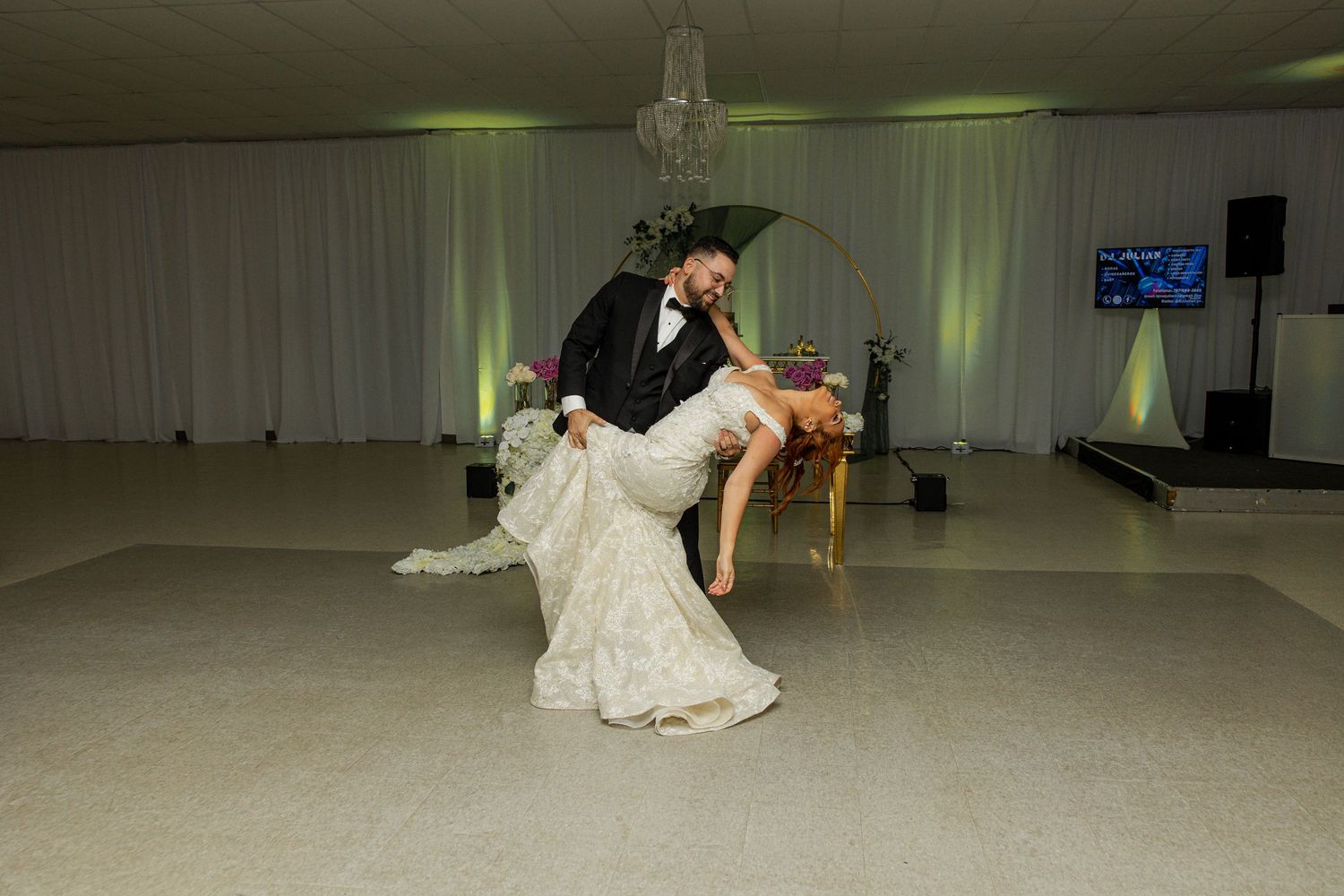 Groom dips bride during romantic first dance on white dance floor at elegant wedding reception.