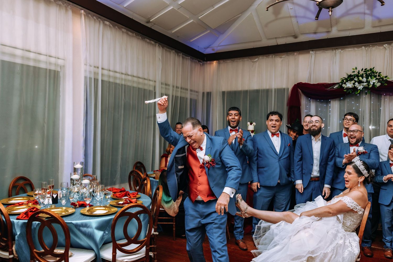 Groom dancing energetically with groomsmen in blue suits and red bow ties at elegant wedding reception.