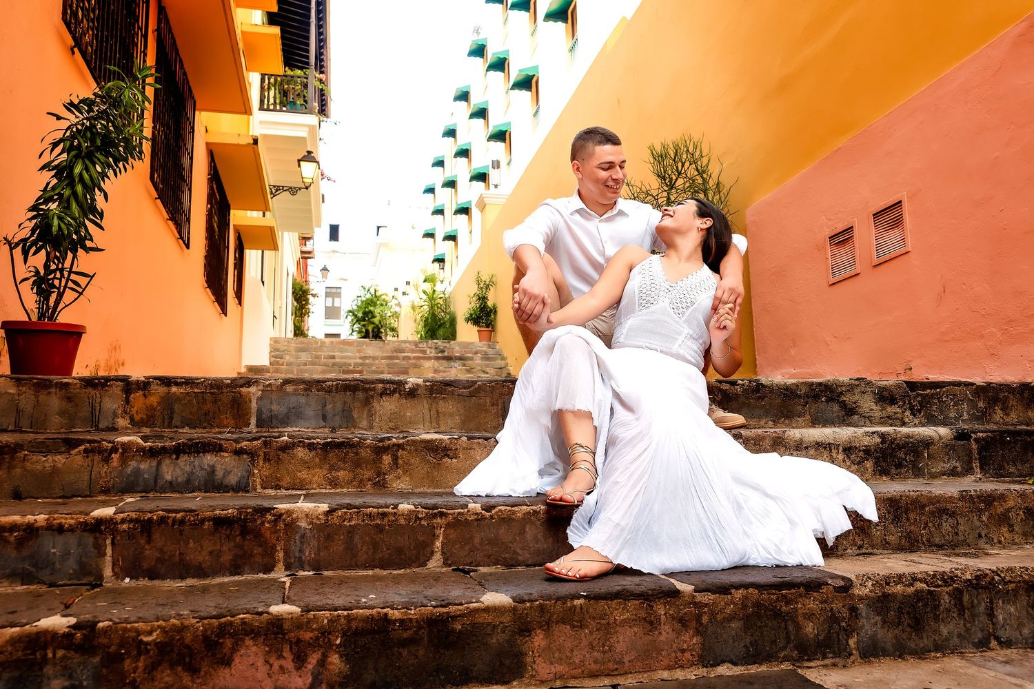 A romantic couple in white outfits sits on rustic stone steps during a tropical engagement photo session.