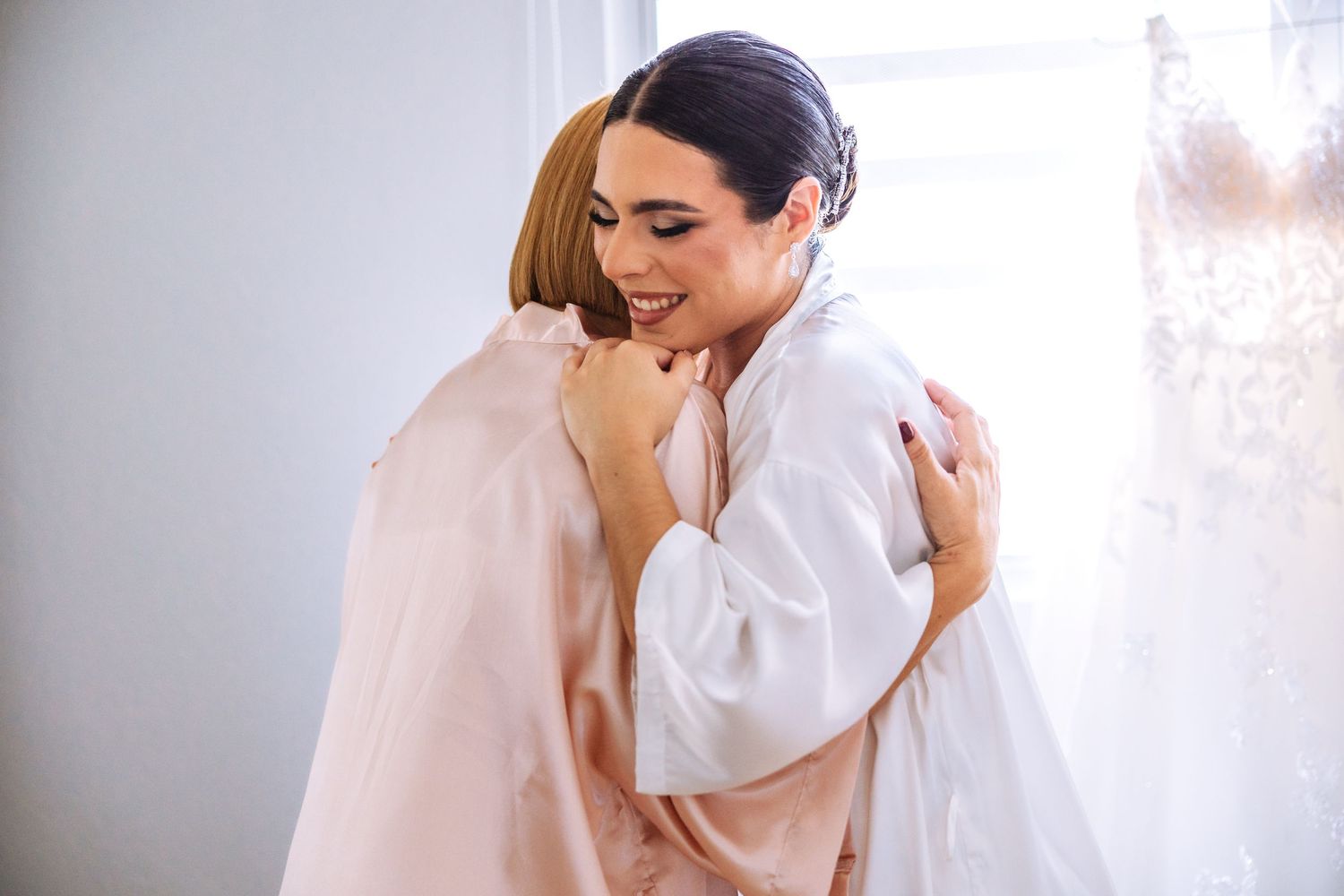 Two people share a warm embrace while wearing light colored clothing in a bright, sunlit room.