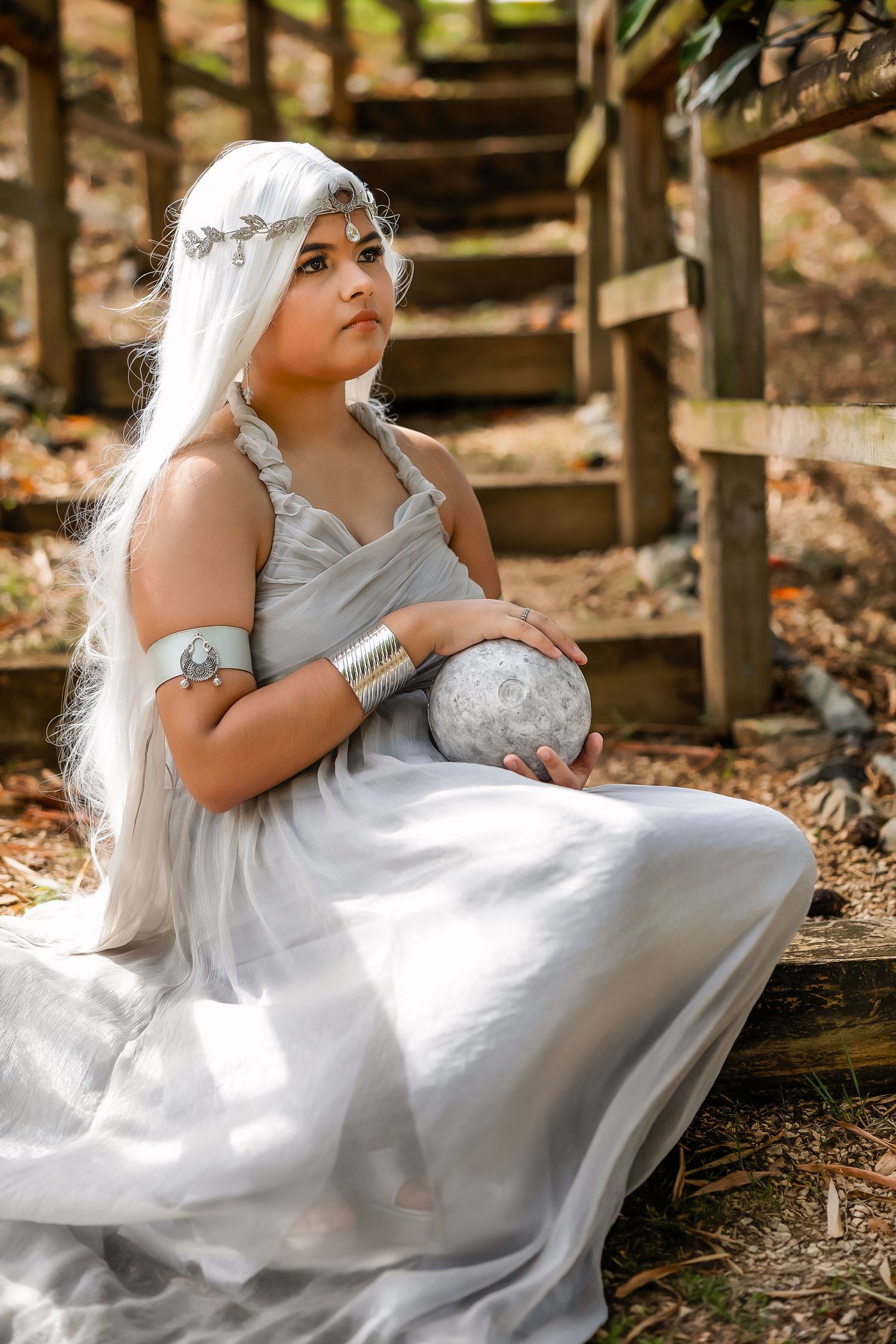 Woman in white gown with silver accessories holds a stone orb while seated on wooden steps in a fantasy cosplay.