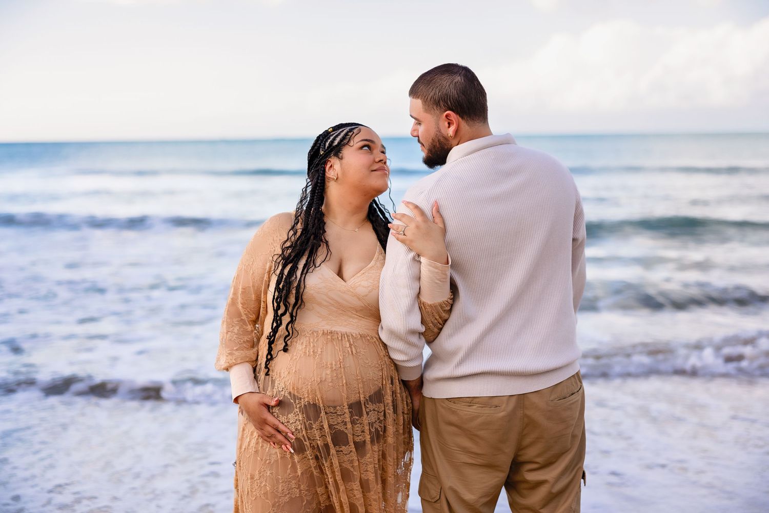 Pregnant couple sharing a tender moment on a beach, woman in beige dress holding baby bump, waves in background.
