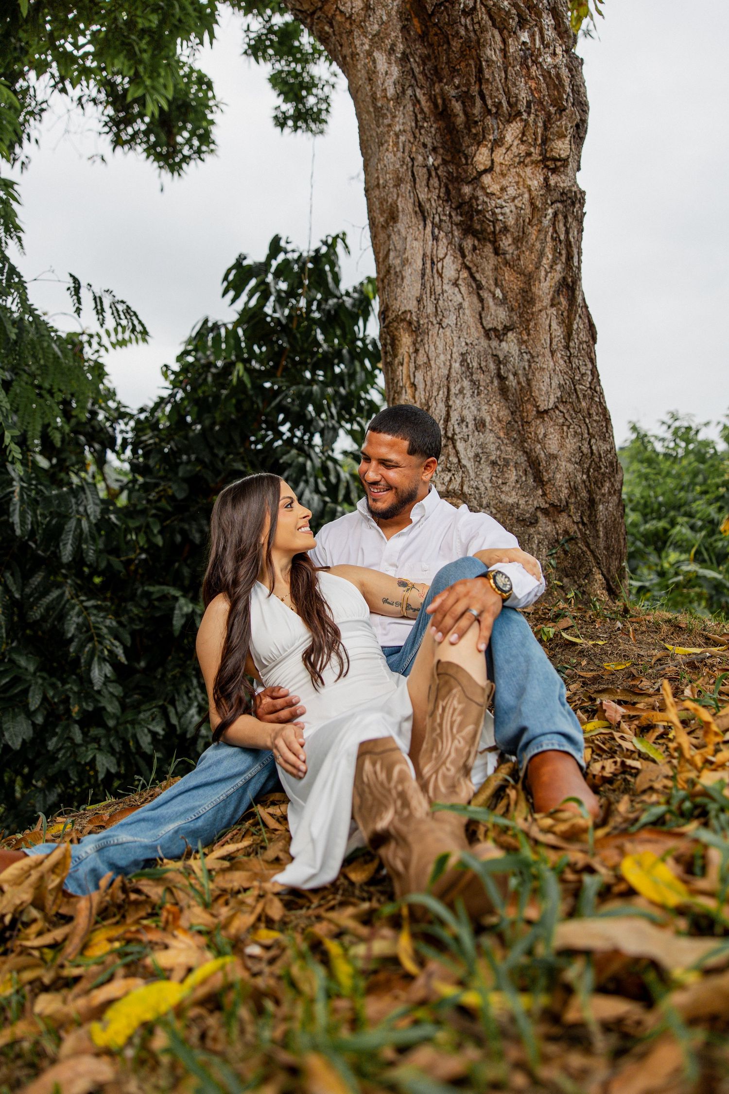Couple sitting together under a large tree surrounded by autumn leaves, woman in white dress and man in white shirt smiling.