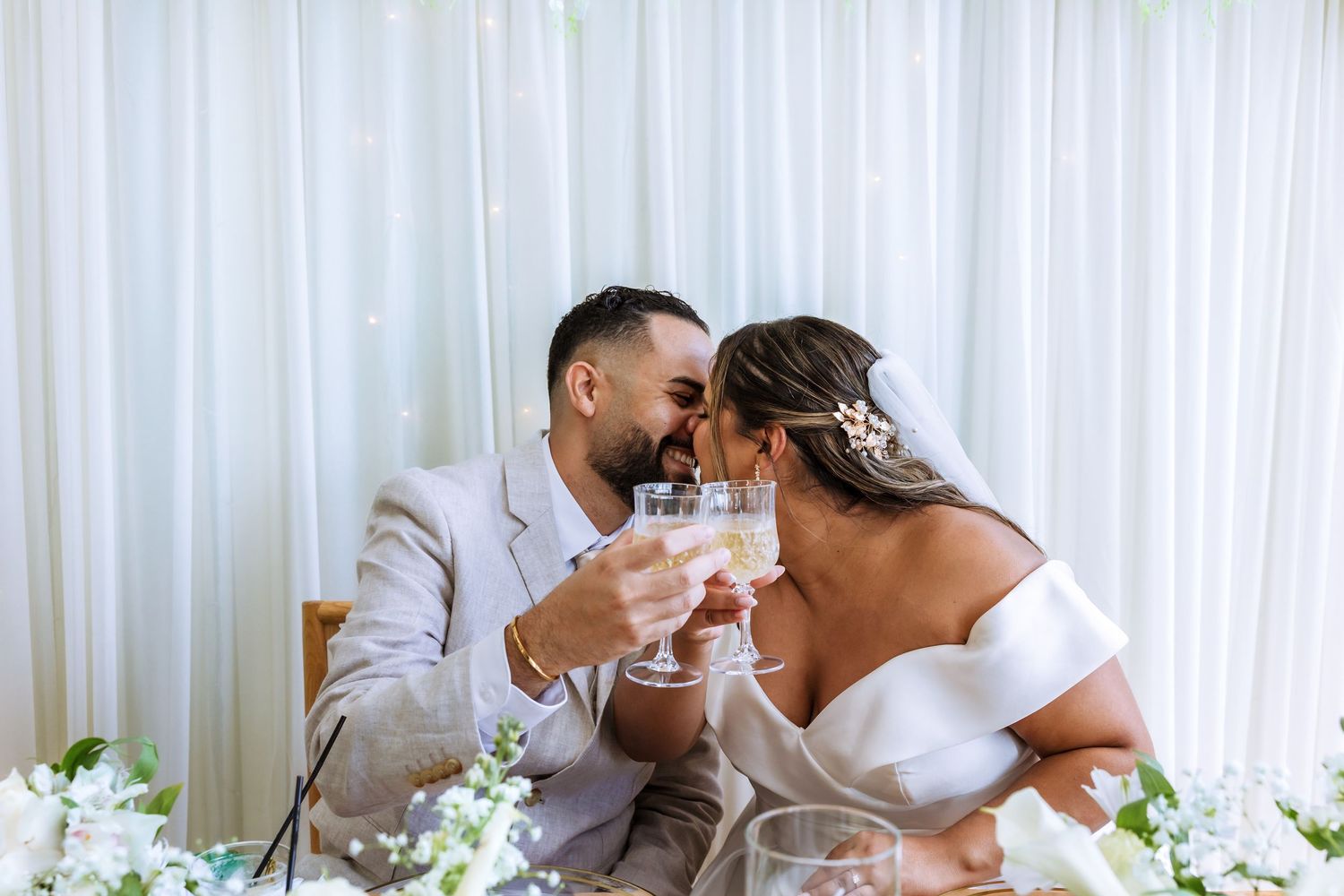 Bride and groom toasting champagne glasses at wedding reception table with white floral decor.