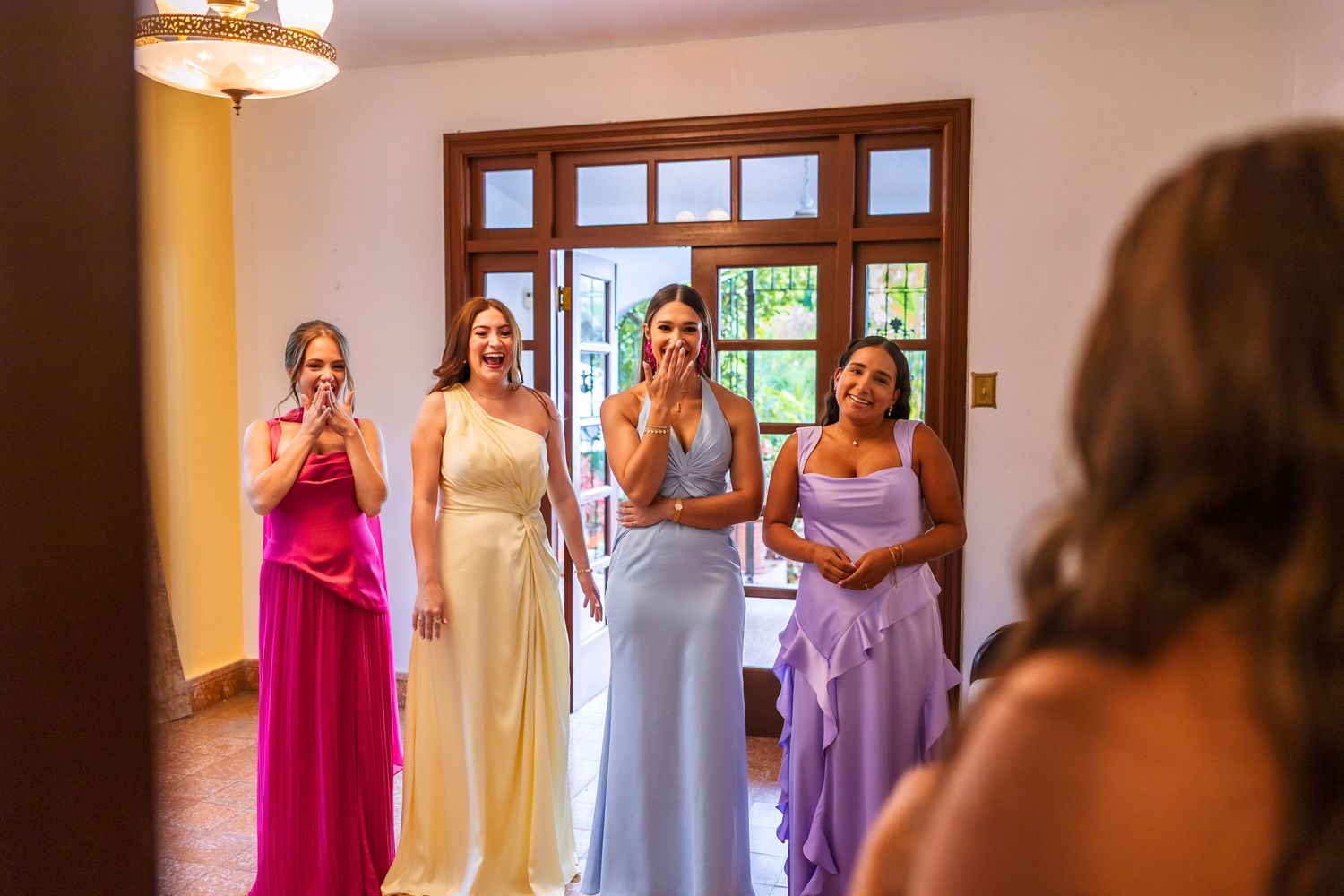 Bridesmaids in colorful gowns react emotionally as bride reveals her wedding dress during bridal preparation.
