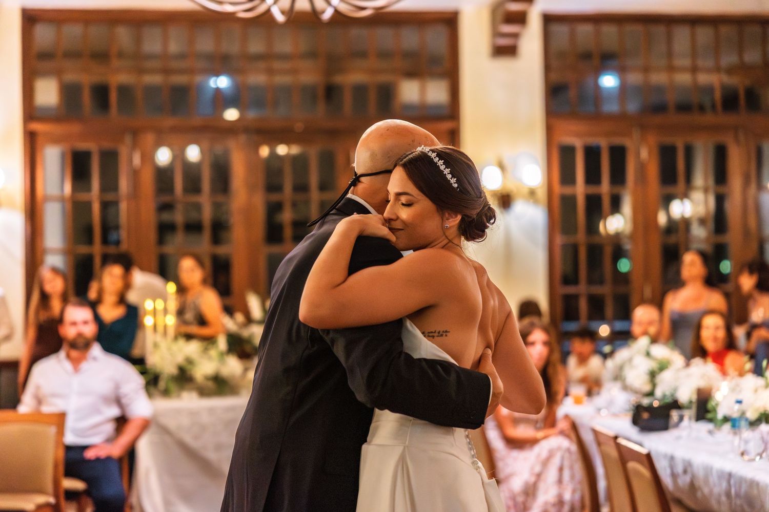 Bride and groom share an emotional first dance embrace at elegant indoor wedding reception with guests seated nearby.