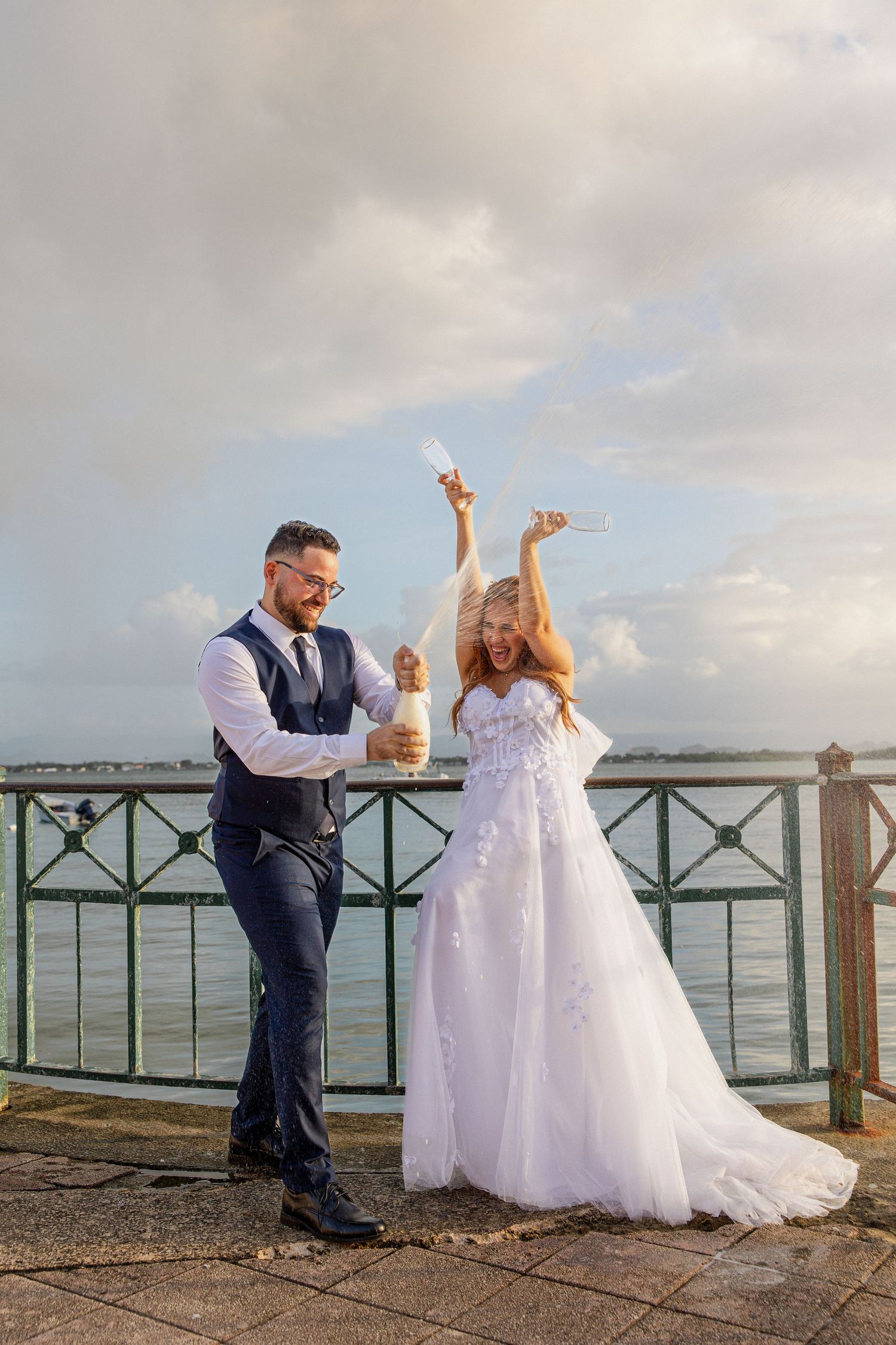Bride and groom celebrating wedding by waterfront, popping champagne bottle with arms raised in joy.