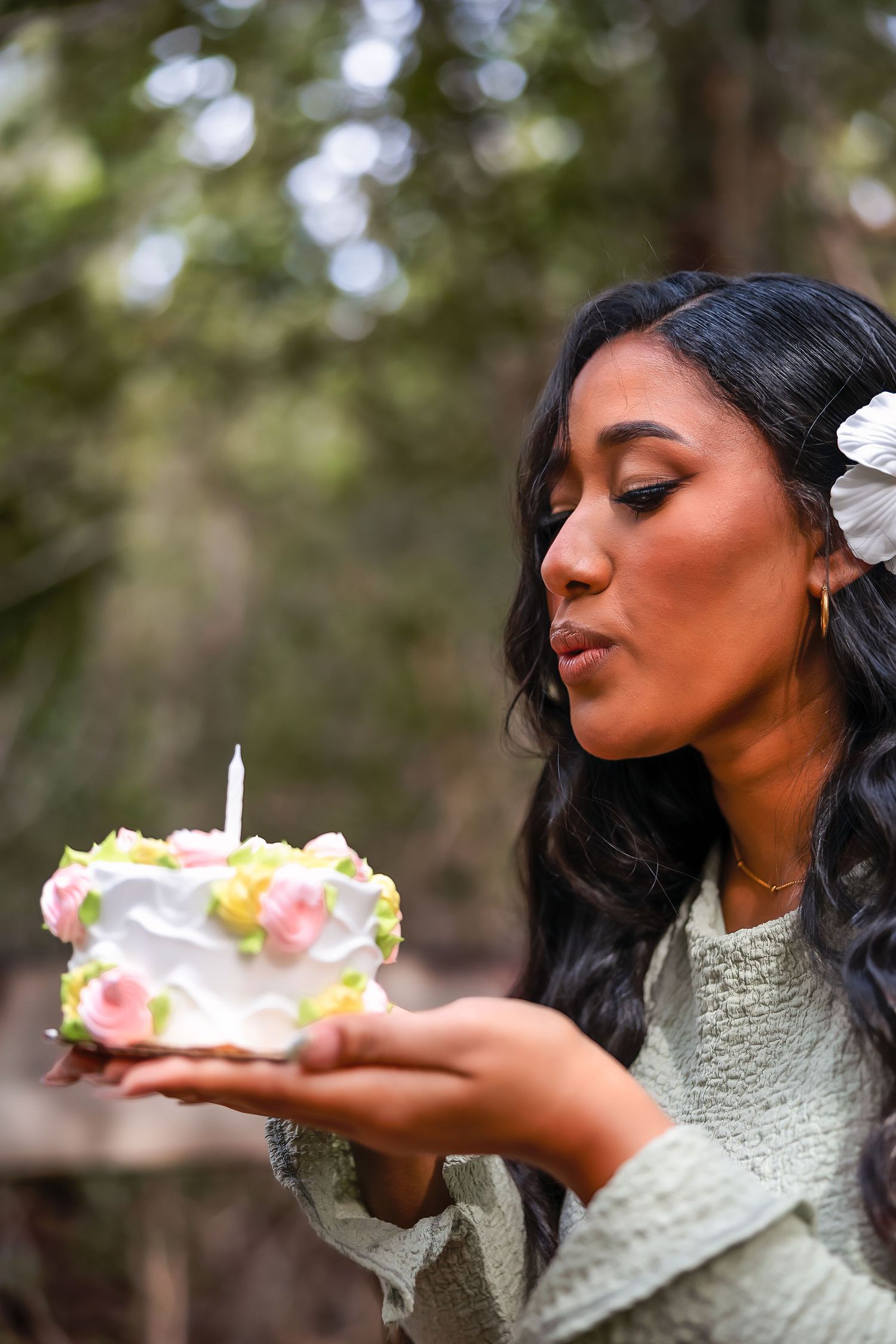Woman in sage green dress blowing out birthday cake candle outdoors, adorned with white flower hair accessory.