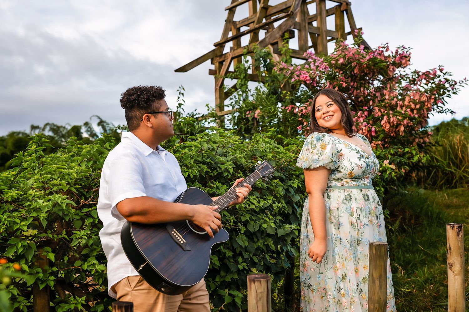 A man with guitar serenades a smiling woman in a floral dress outdoors near blooming roses.