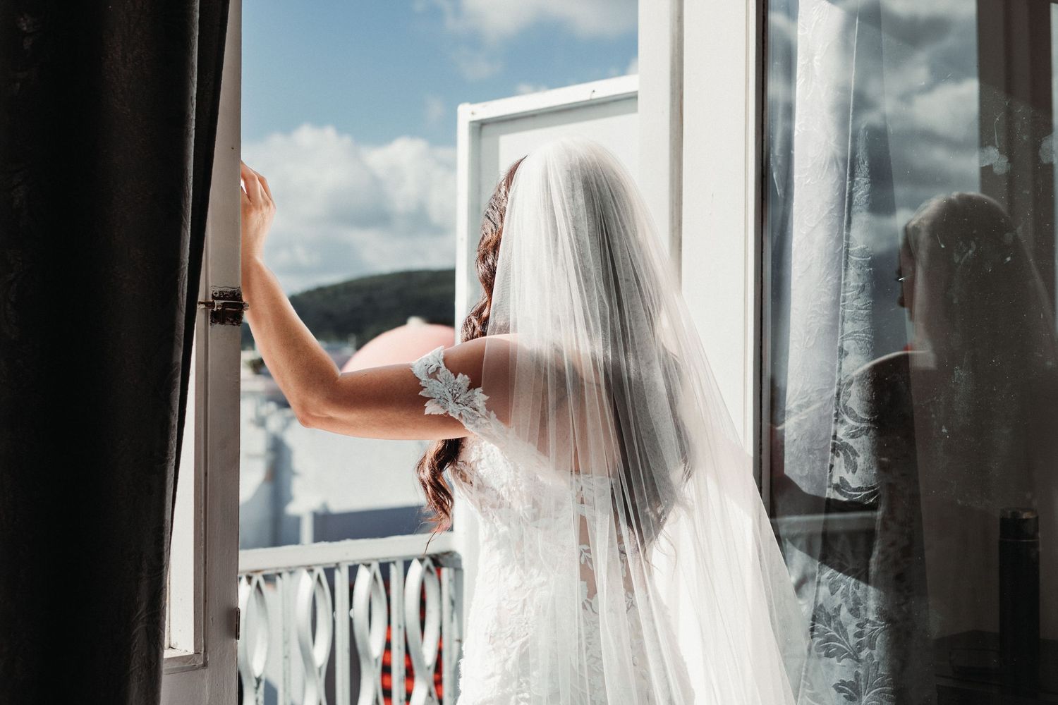 Bride in white lace gown and veil stands at open window, gazing at scenic view on her wedding day.