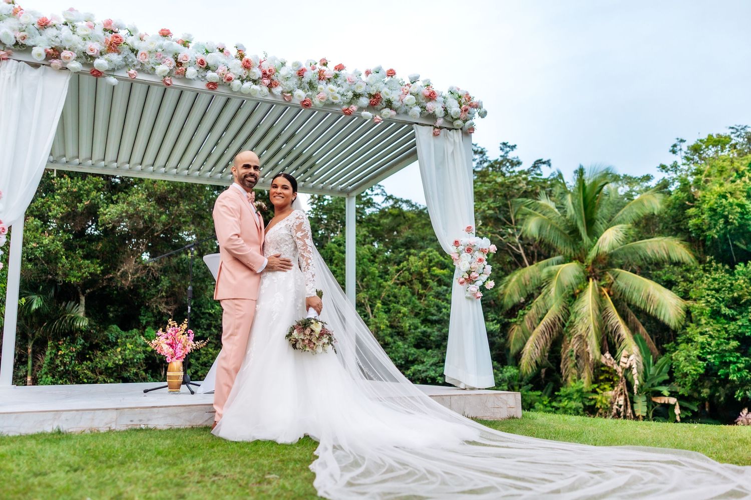 Bride and groom pose under floral arch at outdoor garden wedding ceremony with lush tropical greenery.