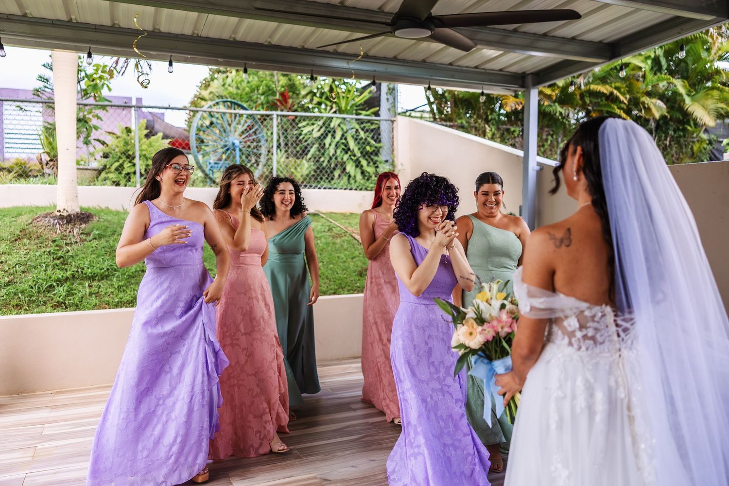 Bridesmaids in colorful mismatched gowns react with joy during a first look moment with a bride in a white wedding dress.
