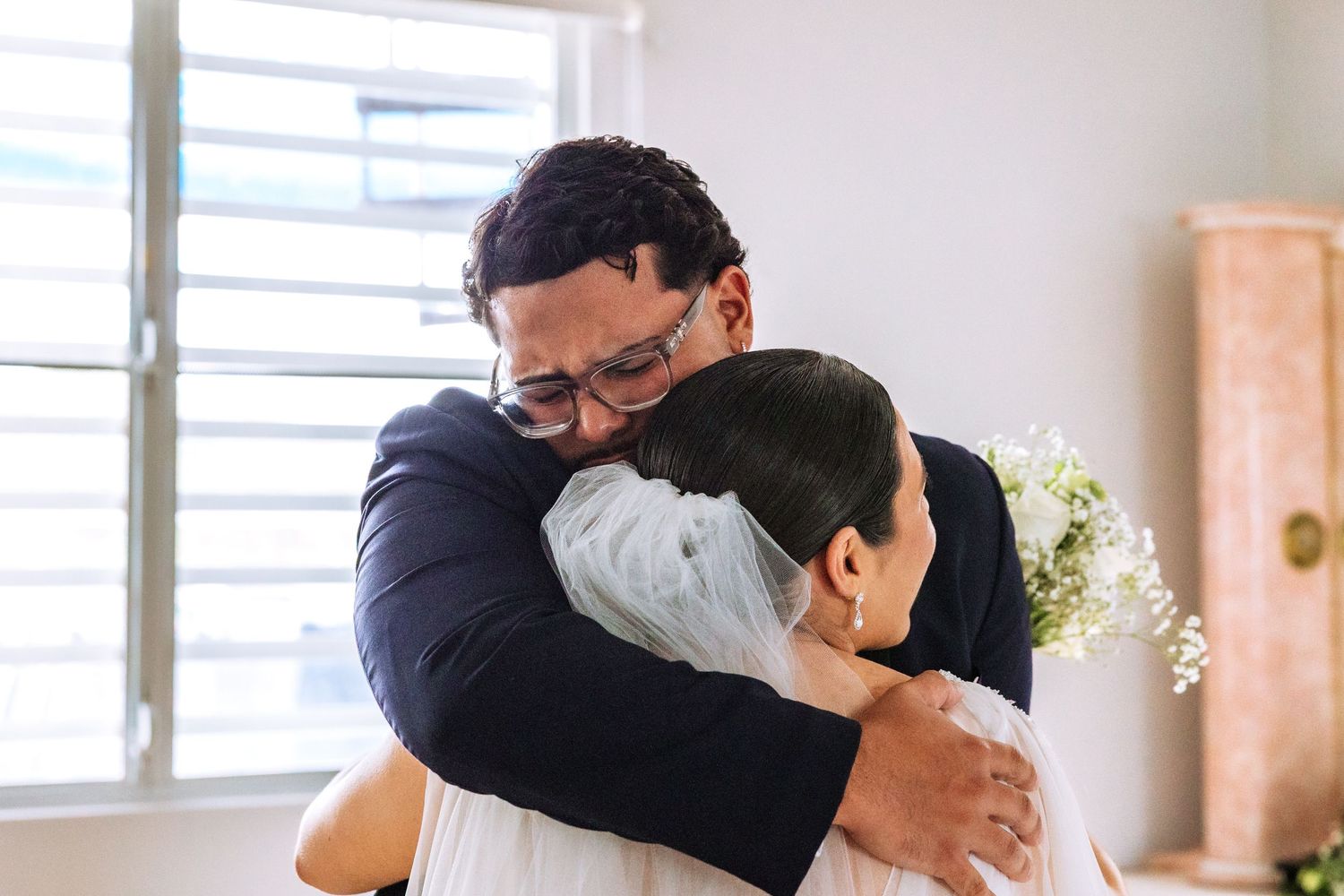 Groom and bride sharing an emotional hug at their wedding ceremony, bride holding small white flowers.
