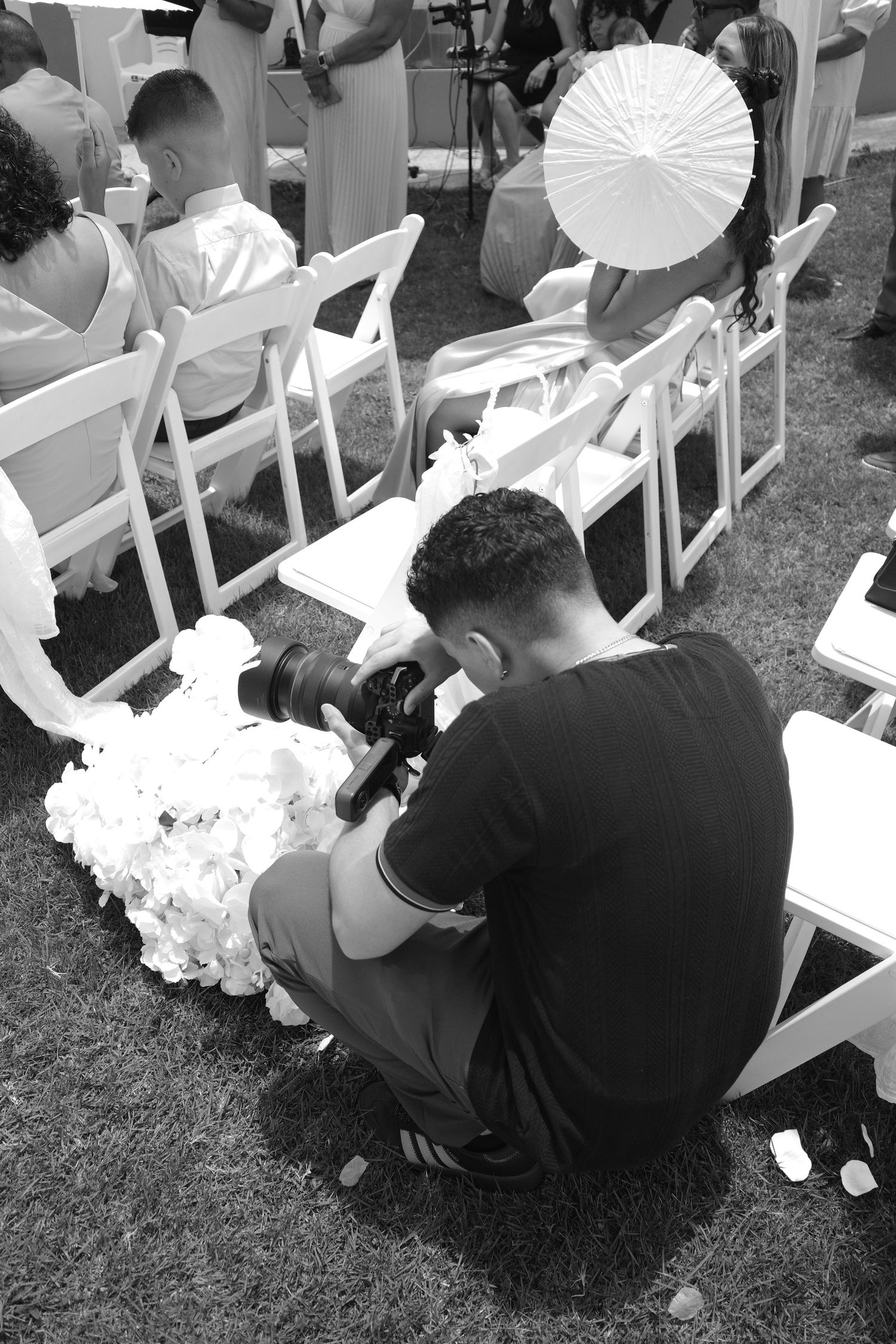 Photographer crouching at outdoor wedding ceremony, capturing floral details with DSLR camera among white chairs.