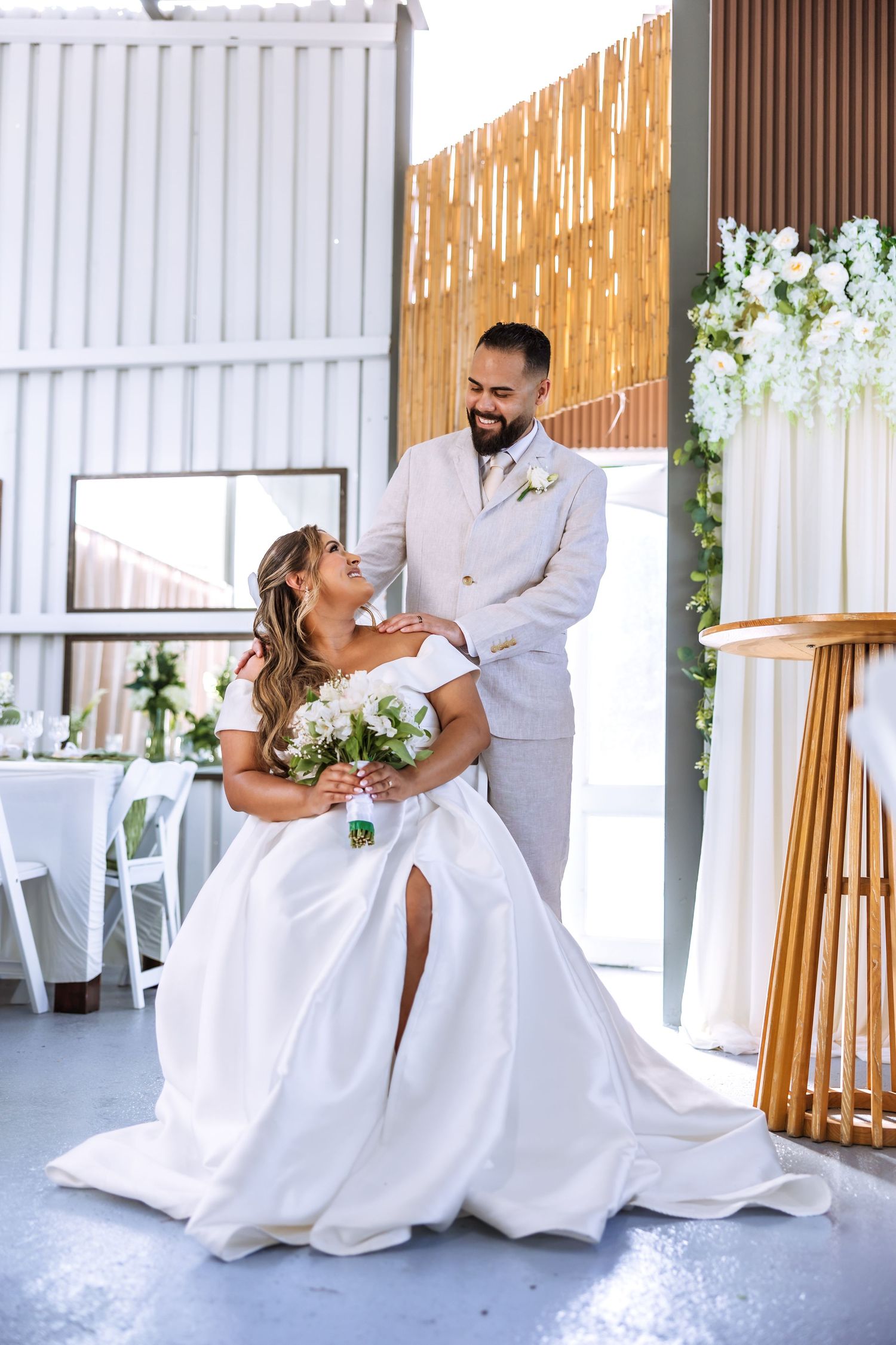 Bride in white gown holding bouquet smiles at groom in beige suit at elegant indoor wedding venue.