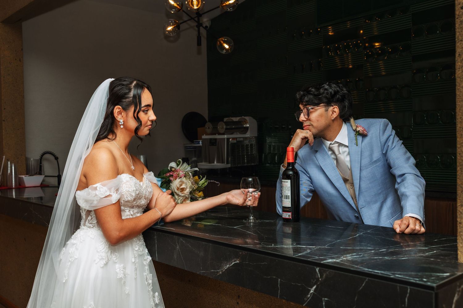 Bride and groom share a romantic moment at a bar, with a wine bottle nearby, bride holding flowers in wedding attire.