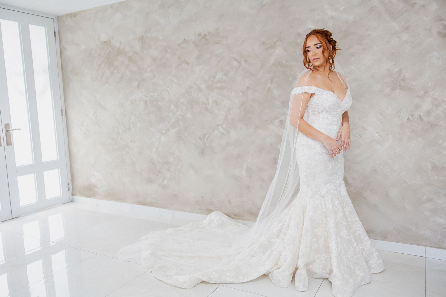Bride in white off-shoulder mermaid wedding gown with lace train and veil poses in elegant bright studio.