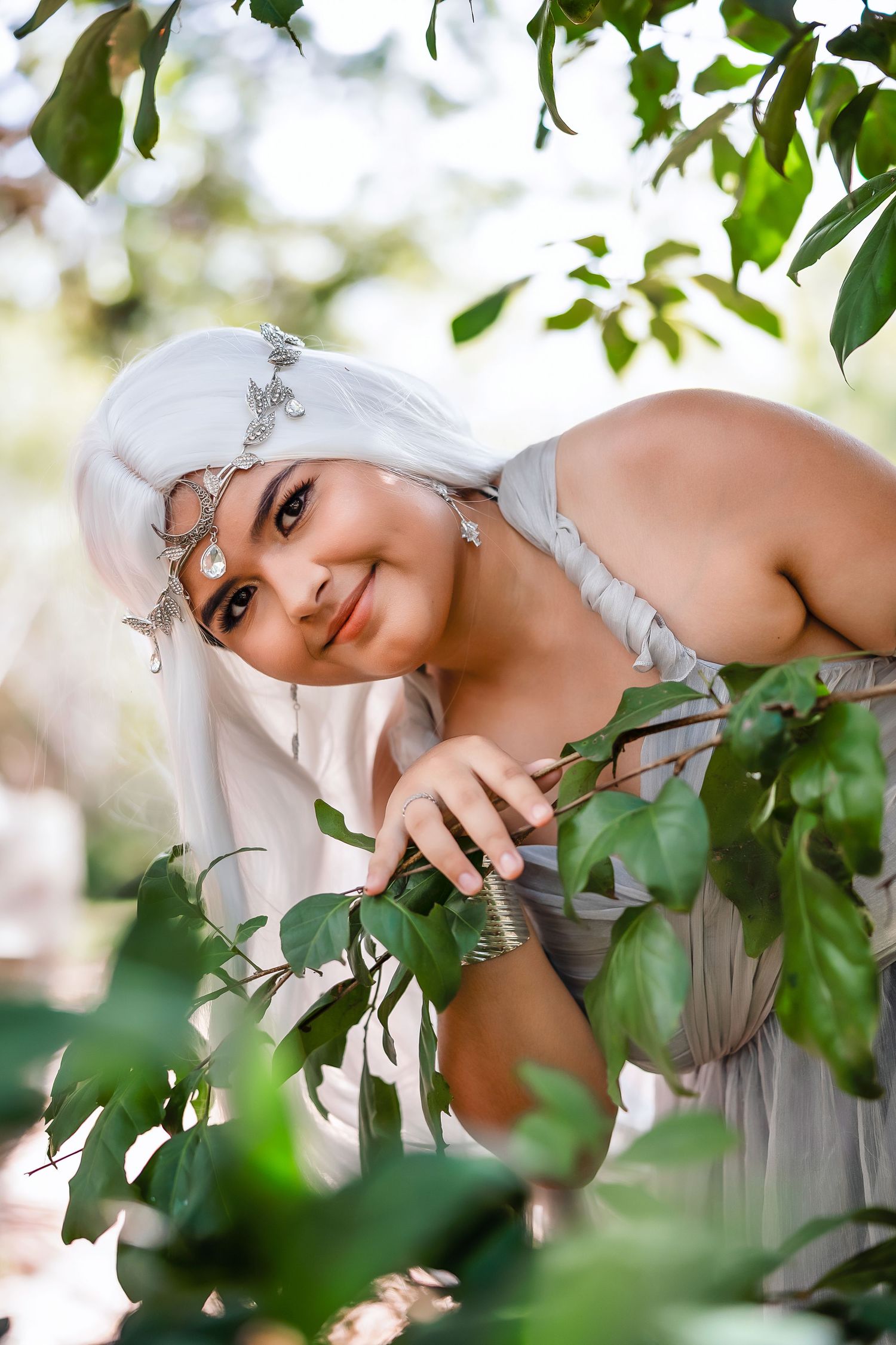 Woman in white hair and silver headpiece poses among green leaves in an ethereal fantasy outdoor portrait.