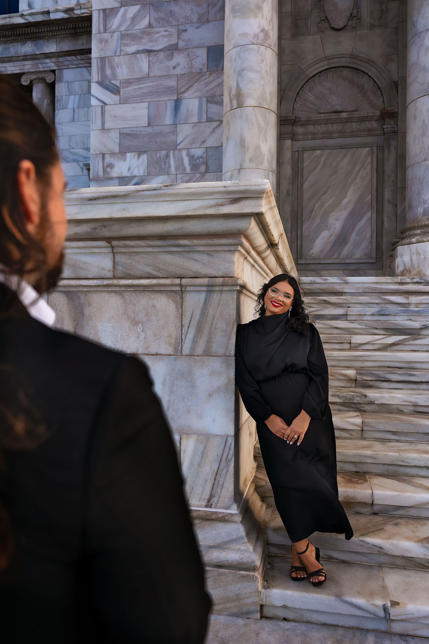 Woman in black dress smiling against marble columns while man in suit stands in foreground.