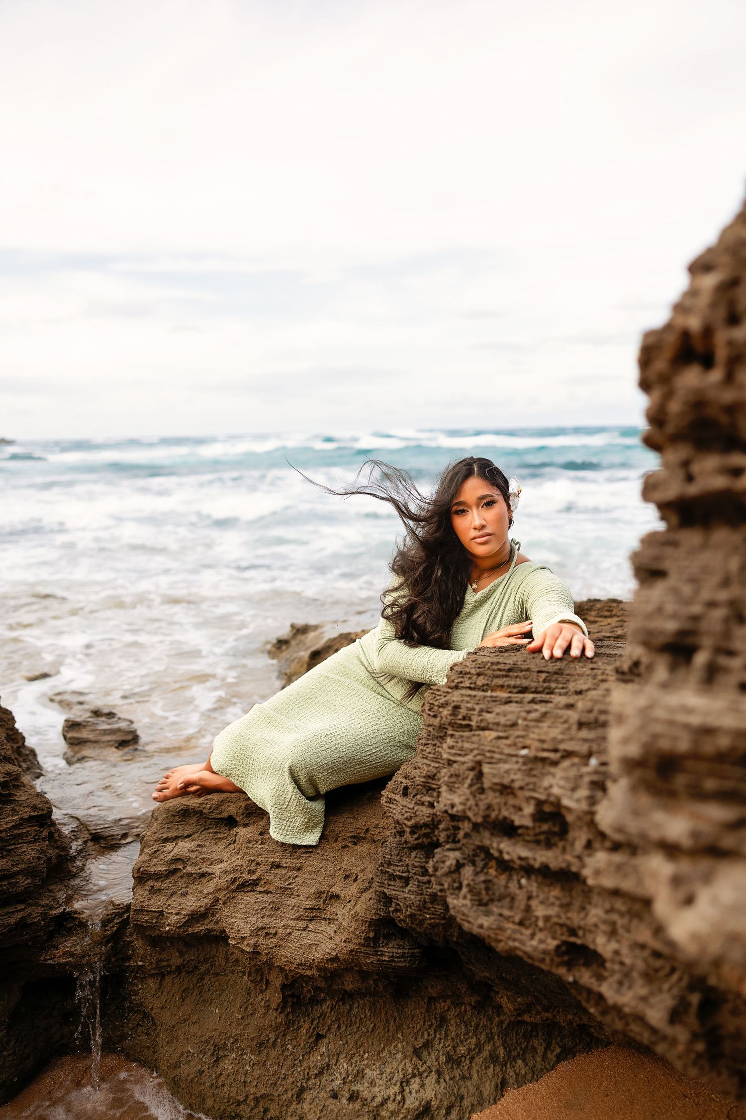 Woman in sage green dress posing on rocky ocean shore with waves and cloudy sky in background.