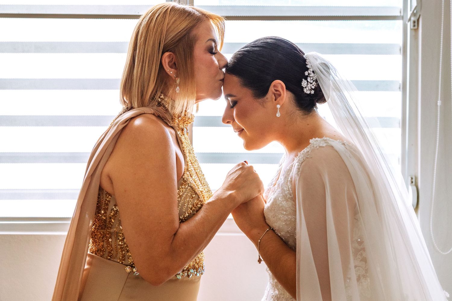 Bride and mother share an intimate forehead-touching moment, holding hands before the wedding ceremony.