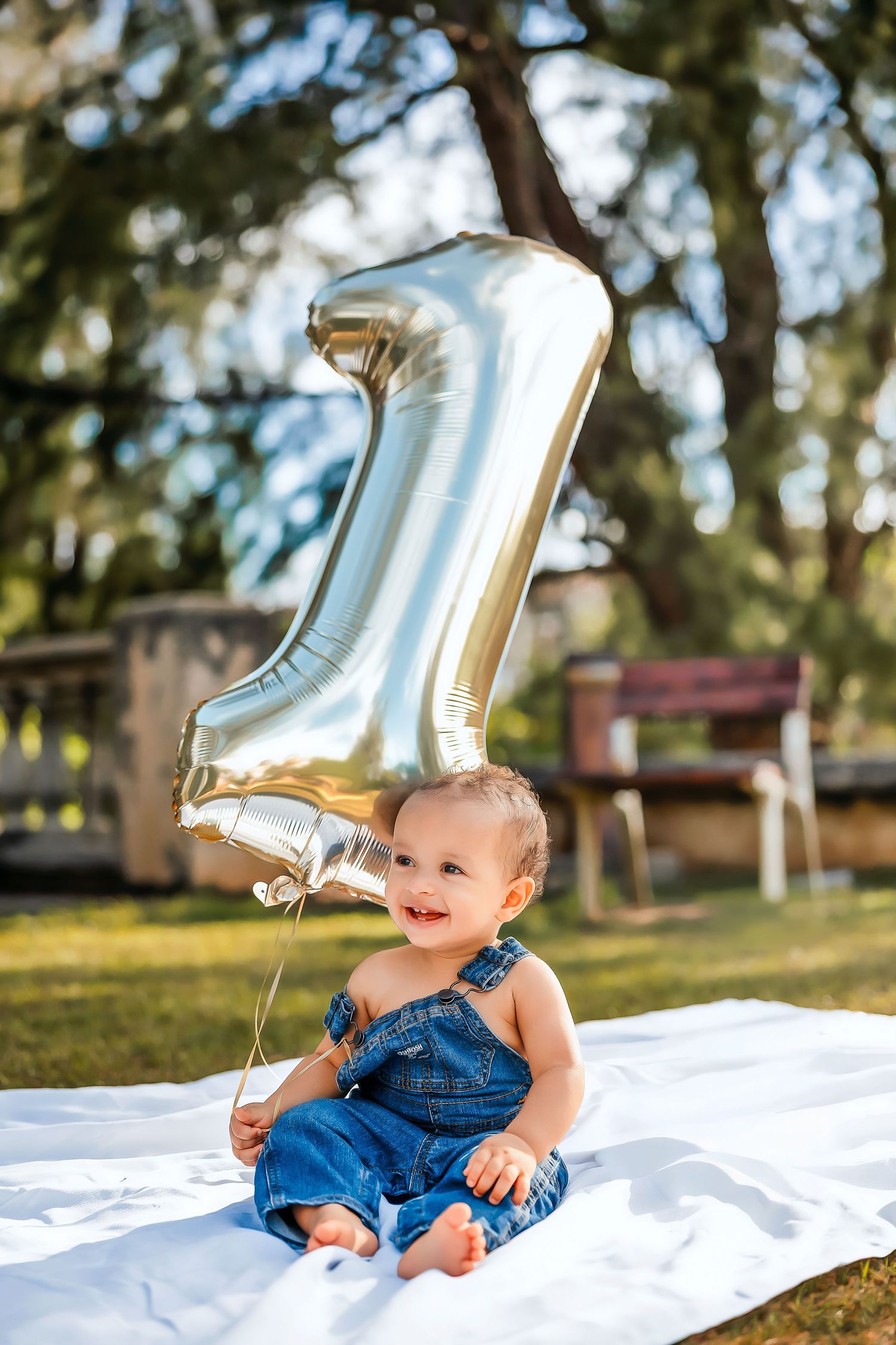 Smiling baby in denim overalls sitting on white blanket outdoors holding a silver number one balloon for first birthday.