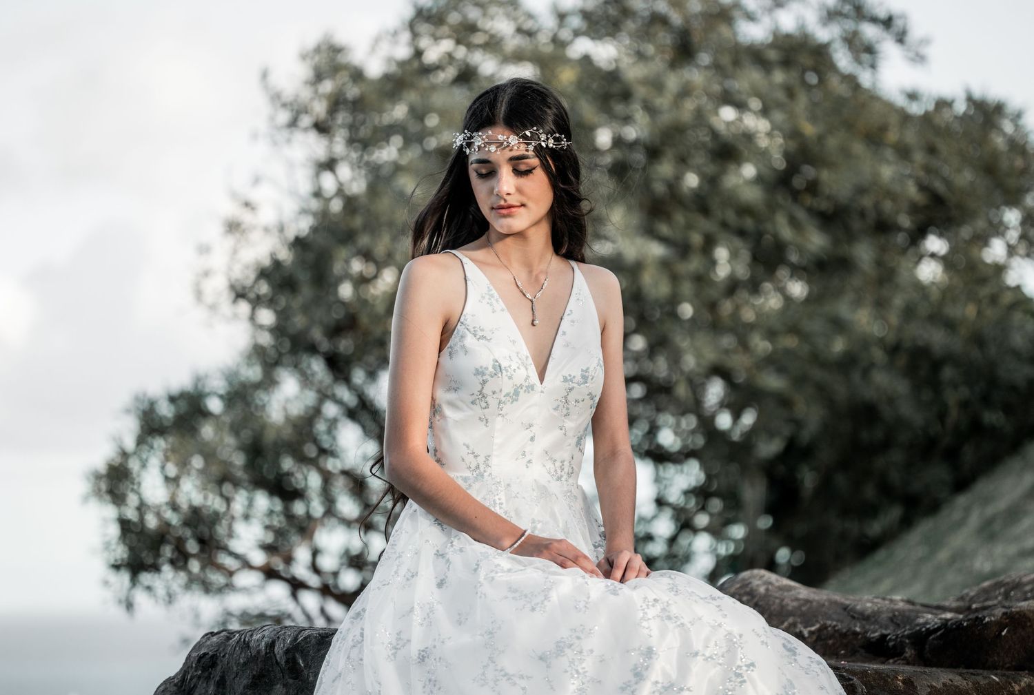 A bride in a white lace V-neck wedding gown with a floral headpiece sits gracefully outdoors.