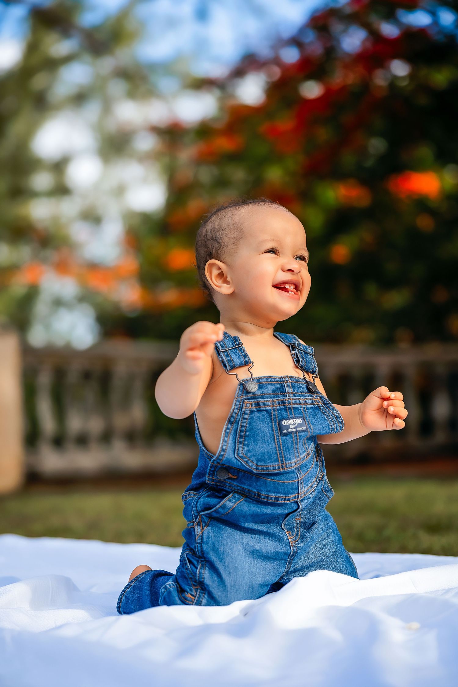 Happy baby in denim overalls sitting on white blanket outdoors, smiling joyfully with colorful garden background.