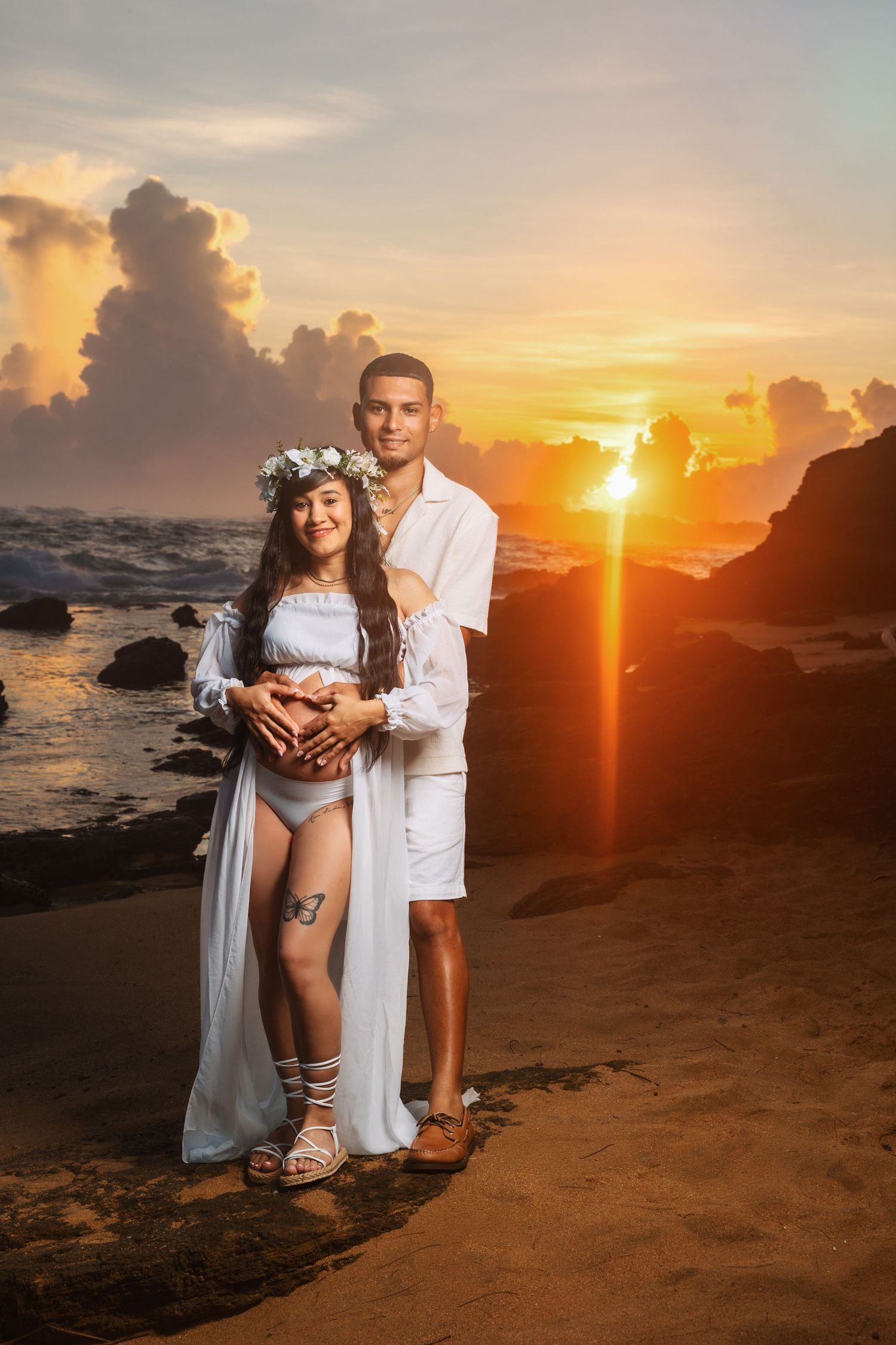 Pregnant couple in white outfits posing on rocky beach at golden sunset, woman wearing floral crown holding baby bump.