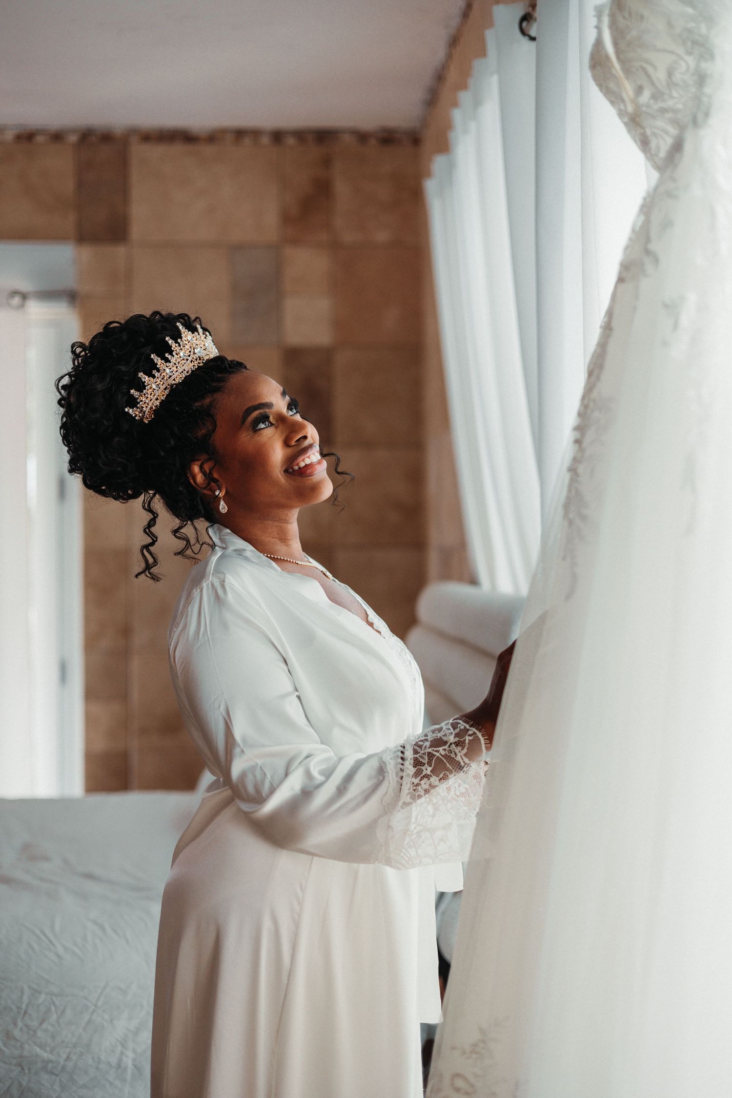 Smiling bride wearing a crown and white robe holds her wedding dress while getting ready in a bright room.