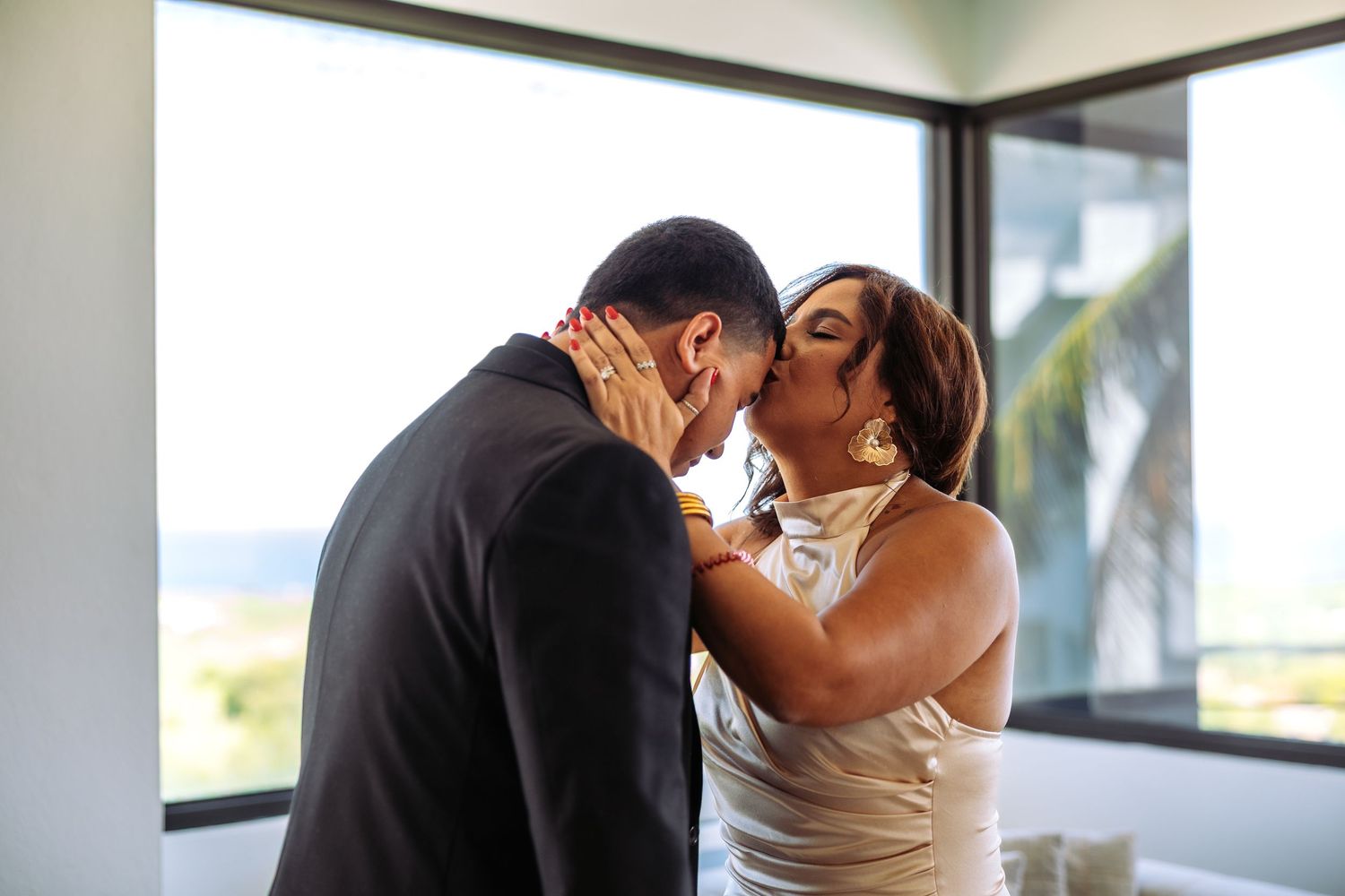 A bride and groom share a romantic kiss near a window with an ocean view on their wedding day.