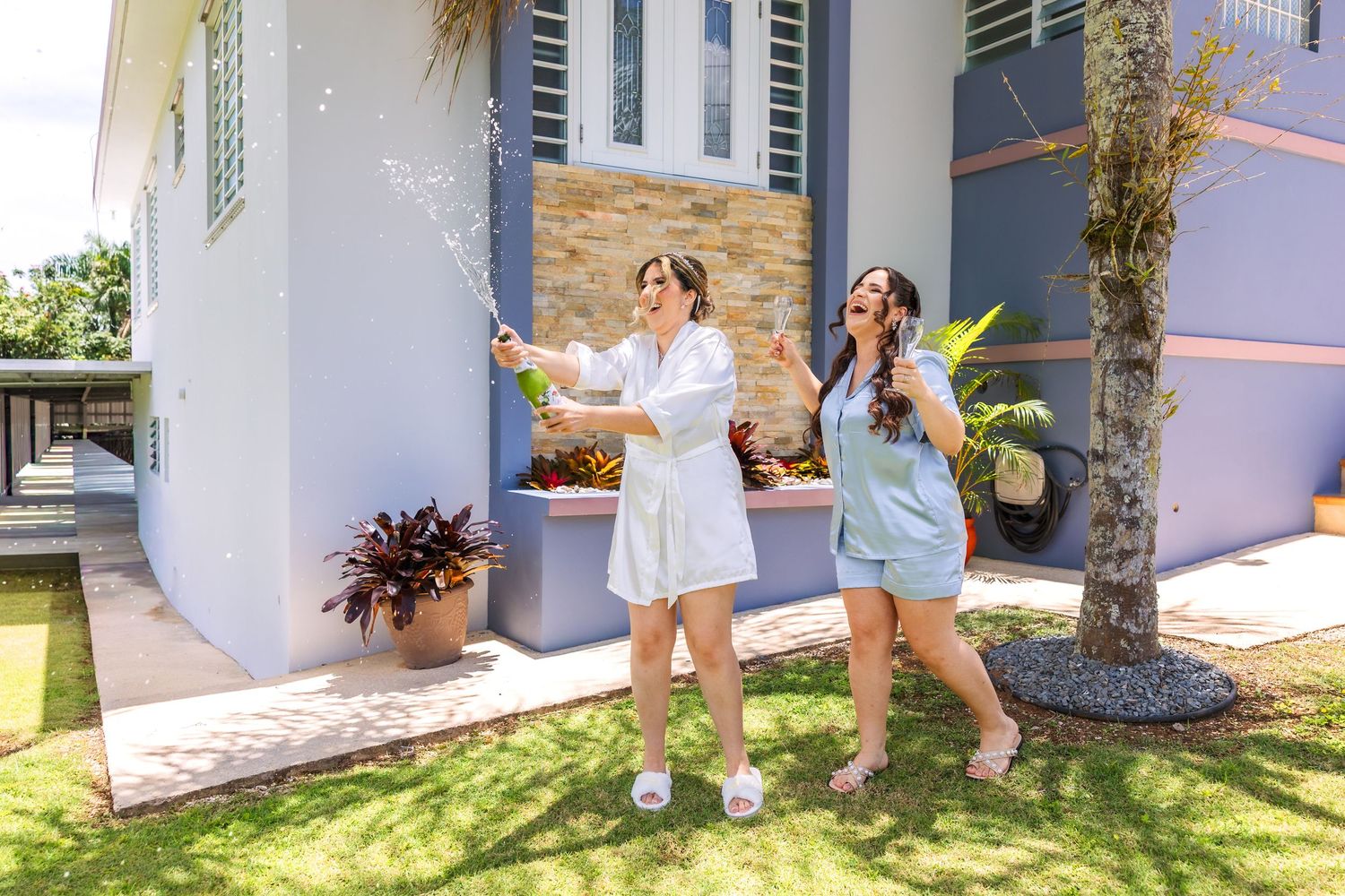 Two women celebrate outdoors, popping champagne in front of a modern home on a sunny day.