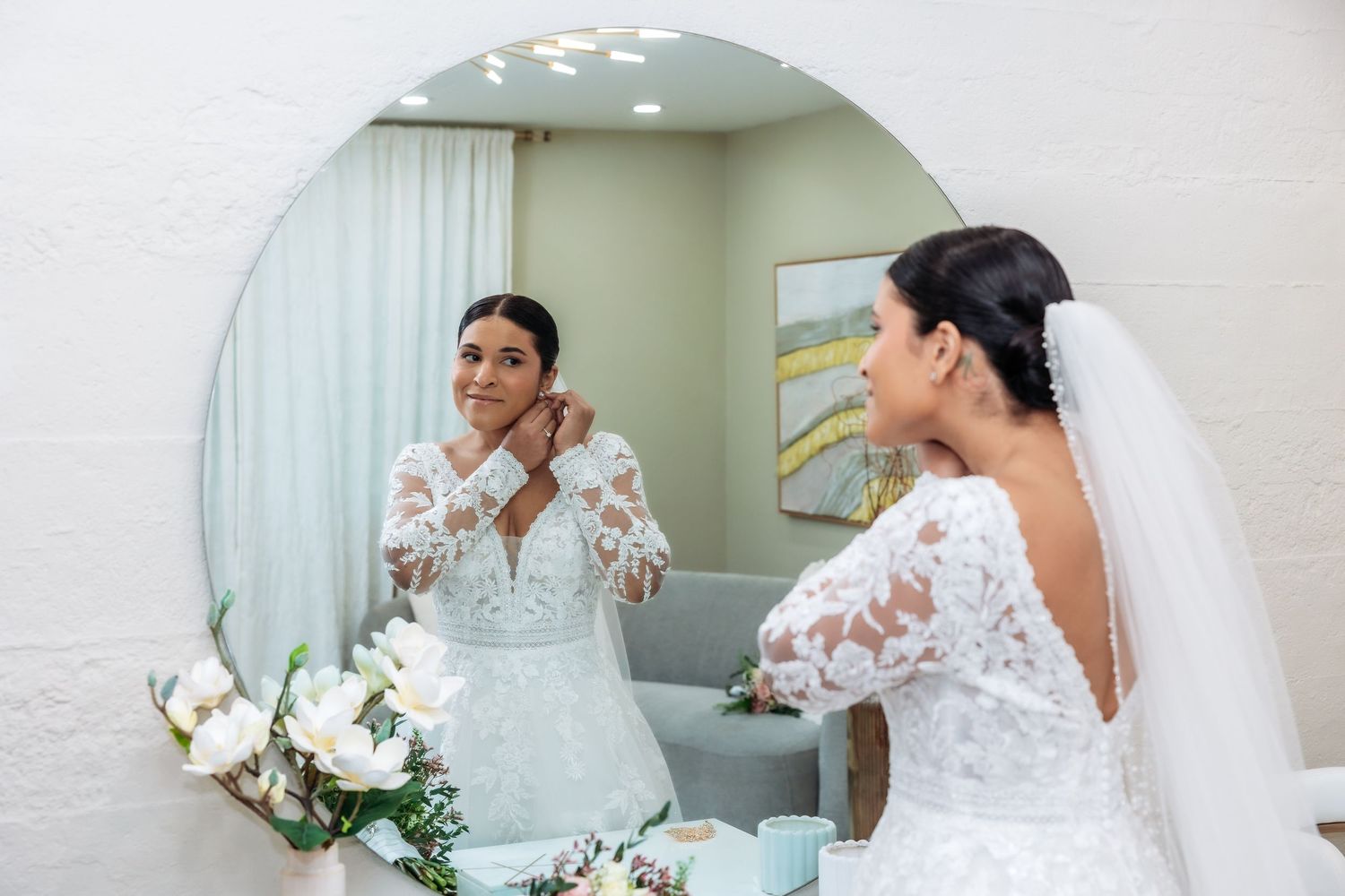 Bride in lace wedding dress adjusting earrings while looking in a round mirror on her wedding day.