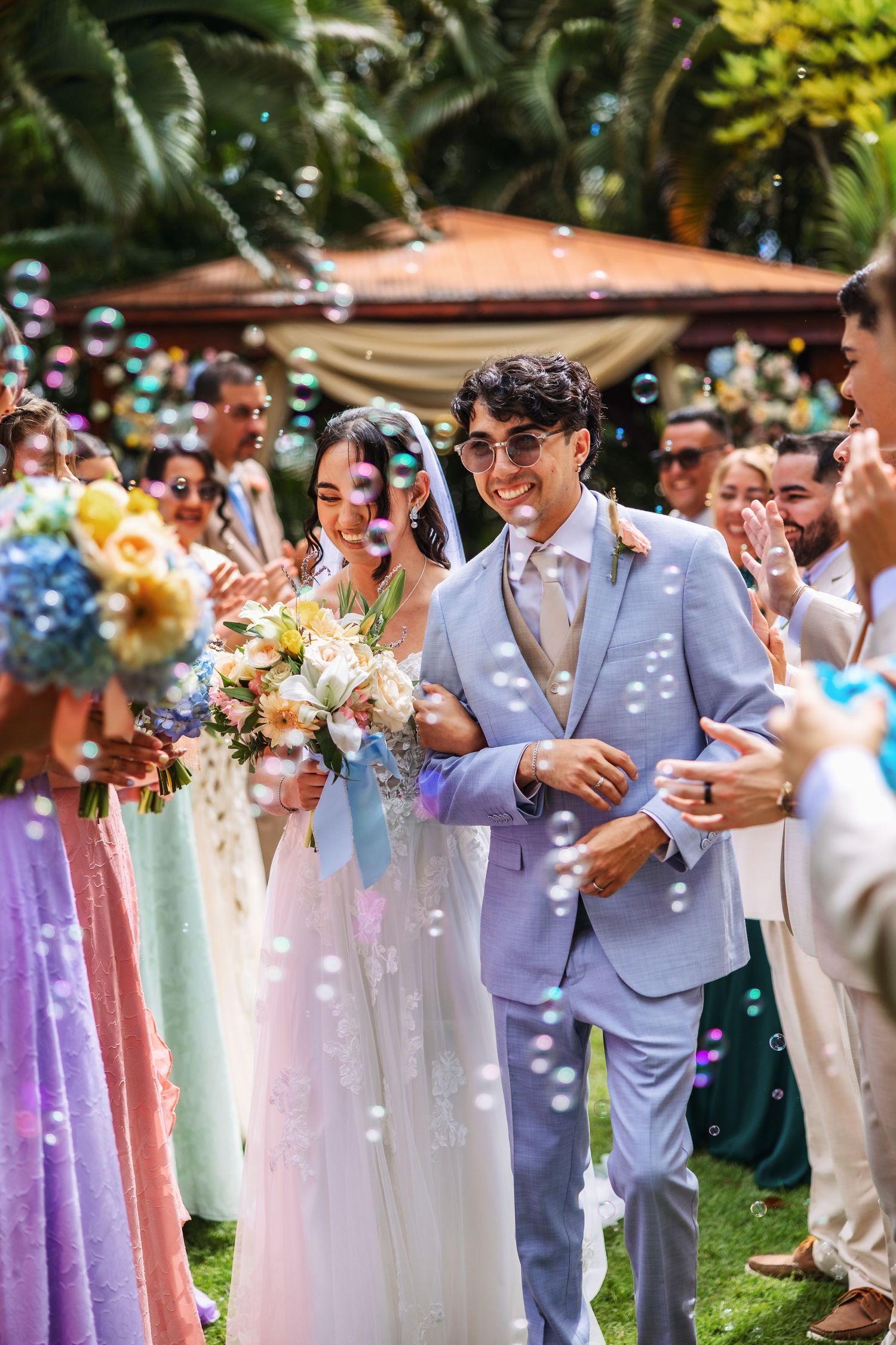 Bride and groom walk through bubble shower at outdoor wedding ceremony surrounded by guests in colorful attire.