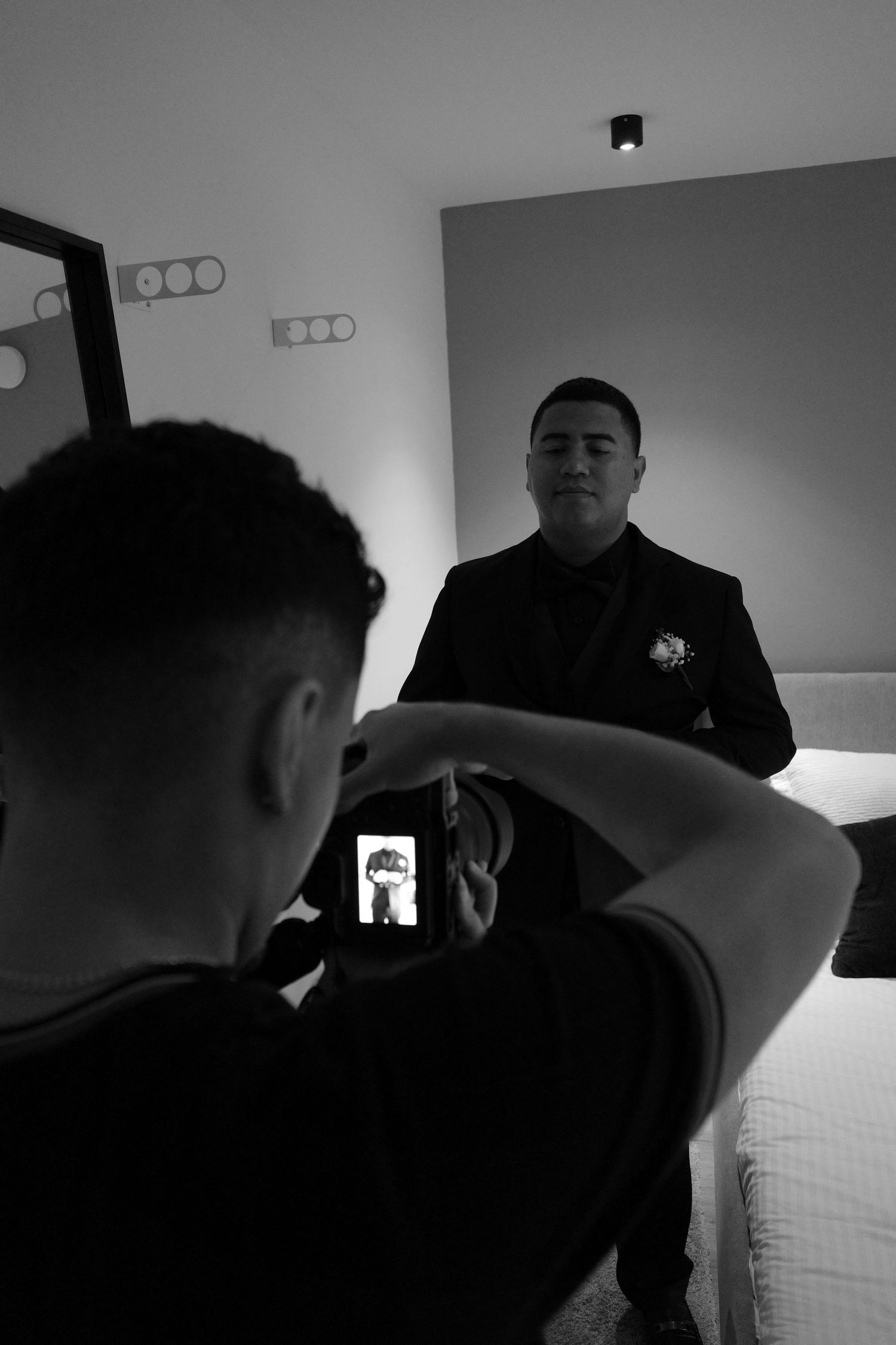 Black and white photo of a man in a suit being photographed by another person holding a smartphone in a hotel room.