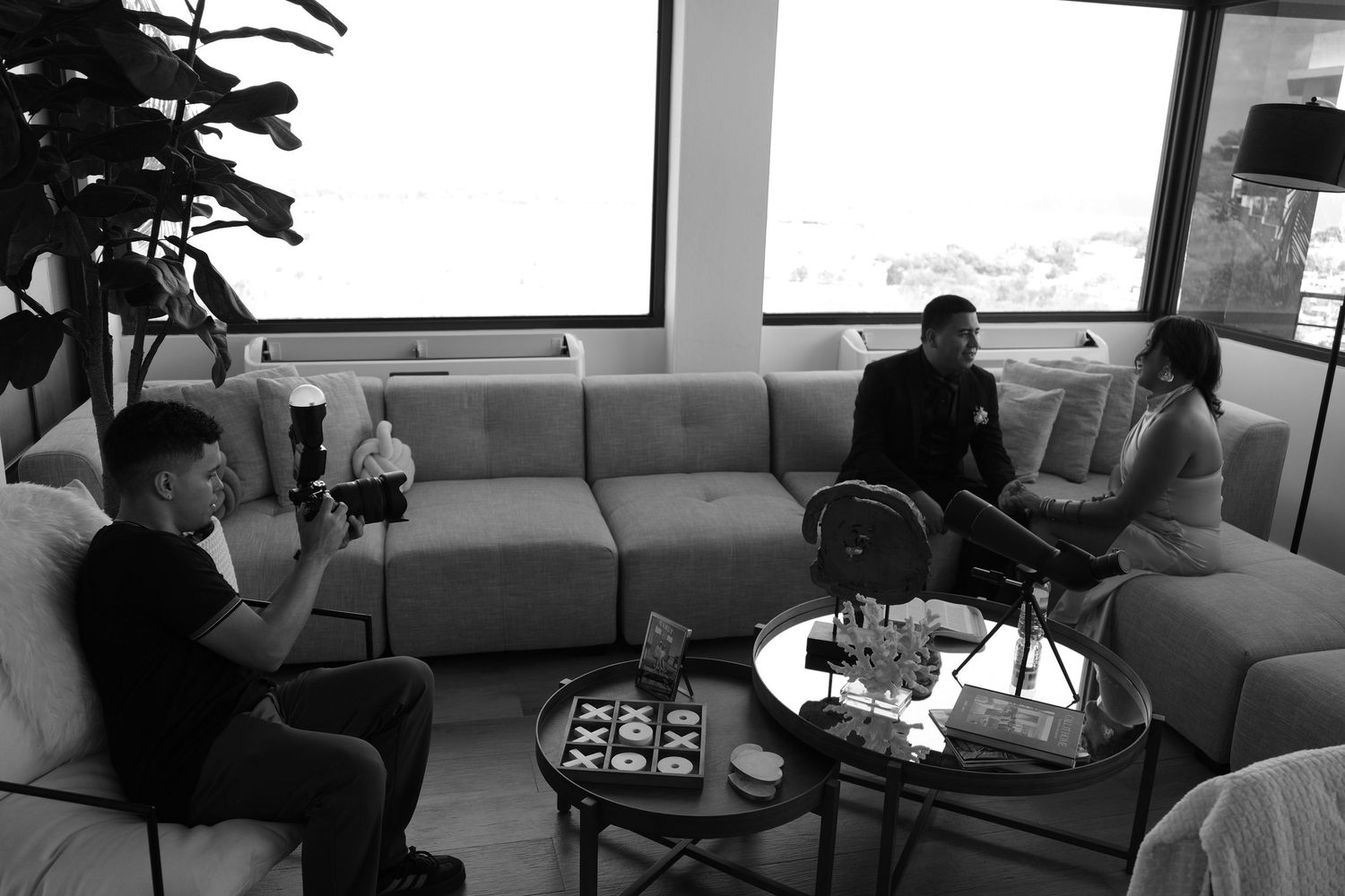 Black and white photo of three people in a modern living room, one filming with a camera during an interview setup.