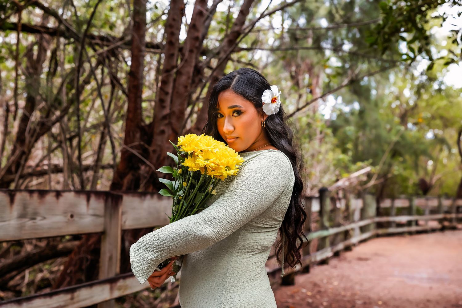 Young woman with flower in hair holds yellow blooms on a wooden bridge path surrounded by trees.
