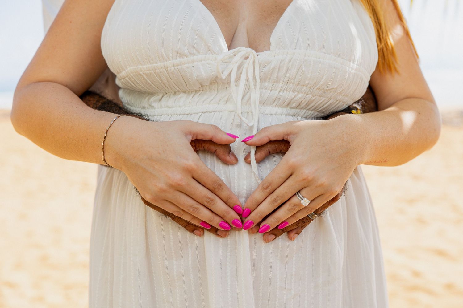 Close-up of hands forming heart shape over white maternity dress.