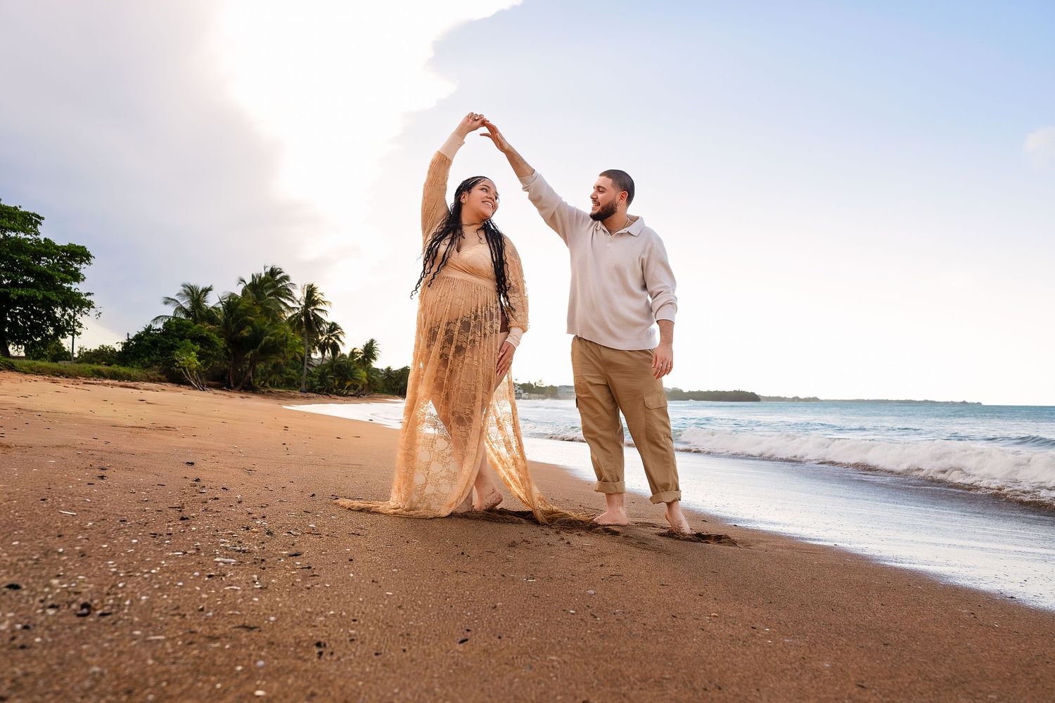Couple dancing on a sandy beach at sunset, woman in a flowing maternity gown.