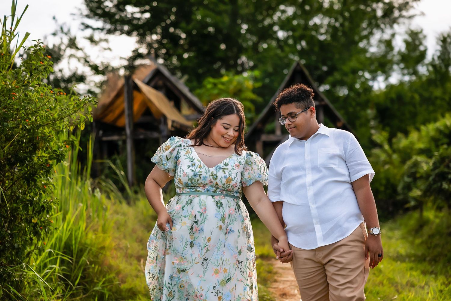 A couple holds hands and walks together through a lush green garden path near a rustic wooden structure.