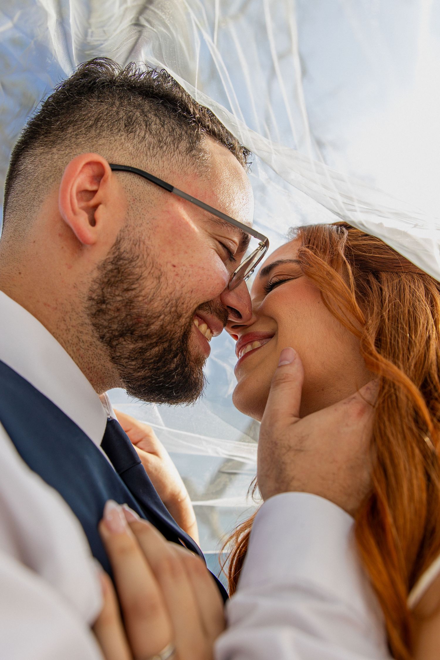 Bride and groom sharing a romantic moment under a wedding veil, smiling and touching foreheads together.