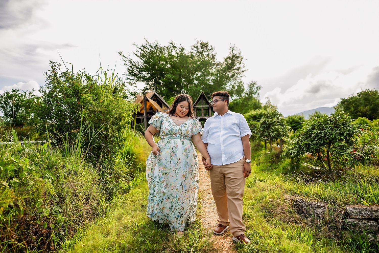 Couple holding hands walking through a lush green garden path near a charming countryside house.